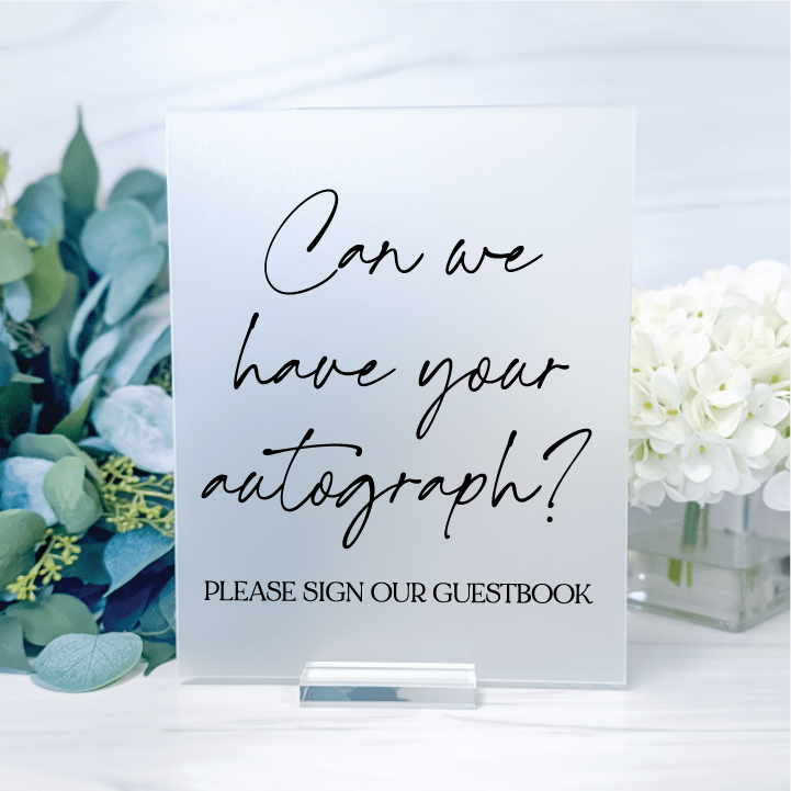 Frosted acrylic wedding guest book sign with black script reading Can We Have Your Autograph, displayed with greenery and white flowers.