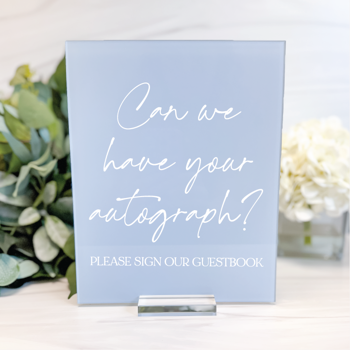 Dusty blue acrylic wedding guest book sign with white handwritten script and Please Sign Our Guestbook wording, styled with wedding greenery.