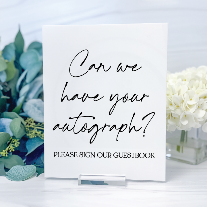 Glossy white acrylic wedding guestbook sign featuring black handwritten lettering and Please Sign Our Guestbook text on a clear display stand.