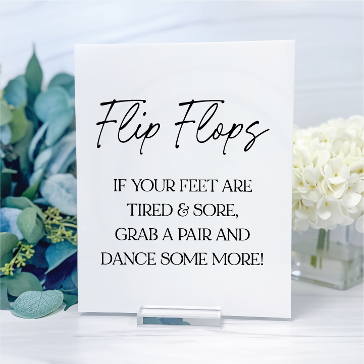 Glossy white rectangular wedding flip flops sign with black handwritten script and dancing shoes poem, displayed on a clear acrylic stand.