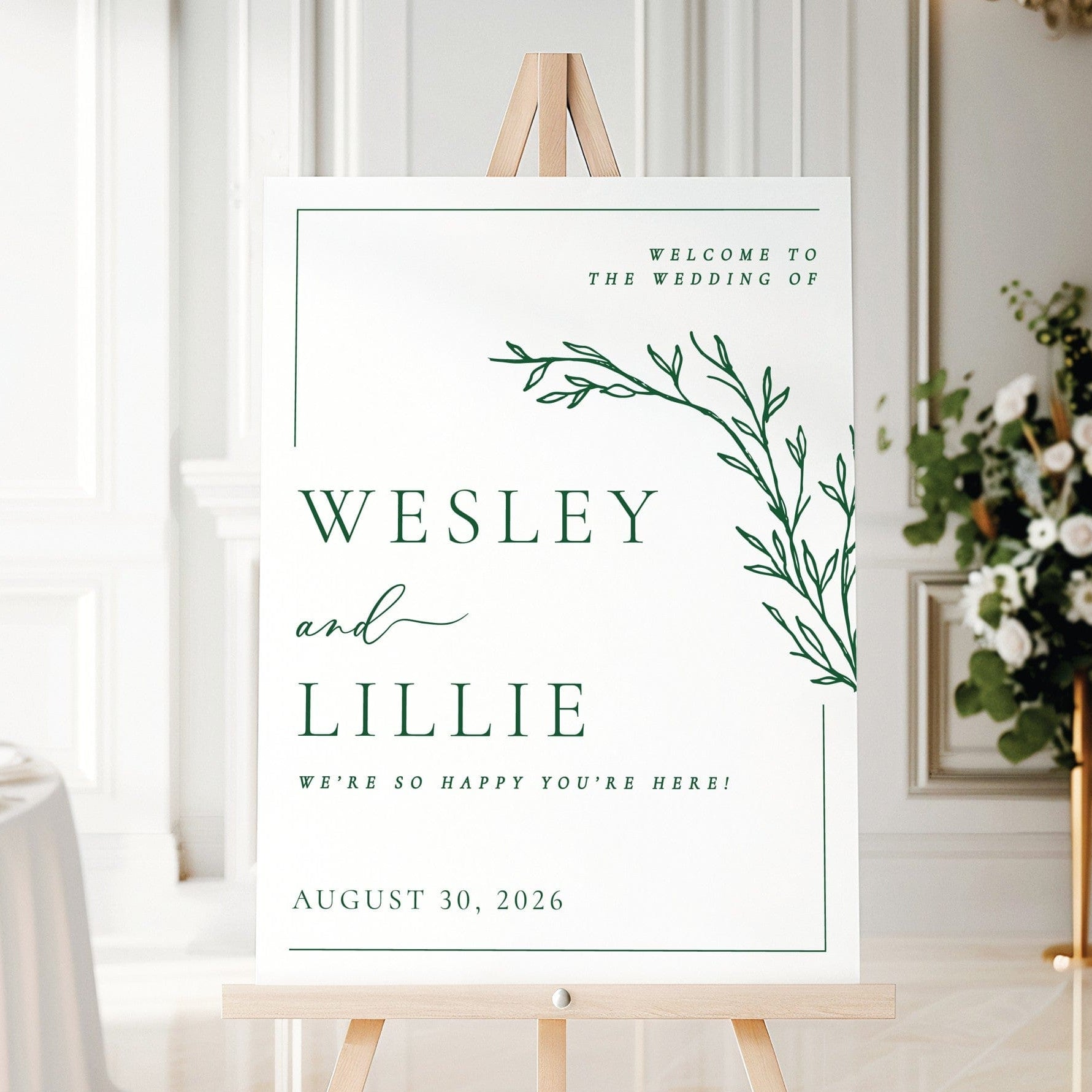 A rectangular white wedding welcome sign features a minimalist green botanical branch design in the top right corner, presented on a light wood easel.