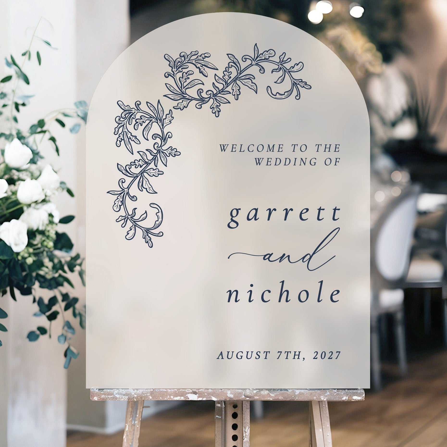 An elegant arched frosted welcome sign for a wedding, featuring intricate dark blue floral scrollwork and displayed on a wooden easel.