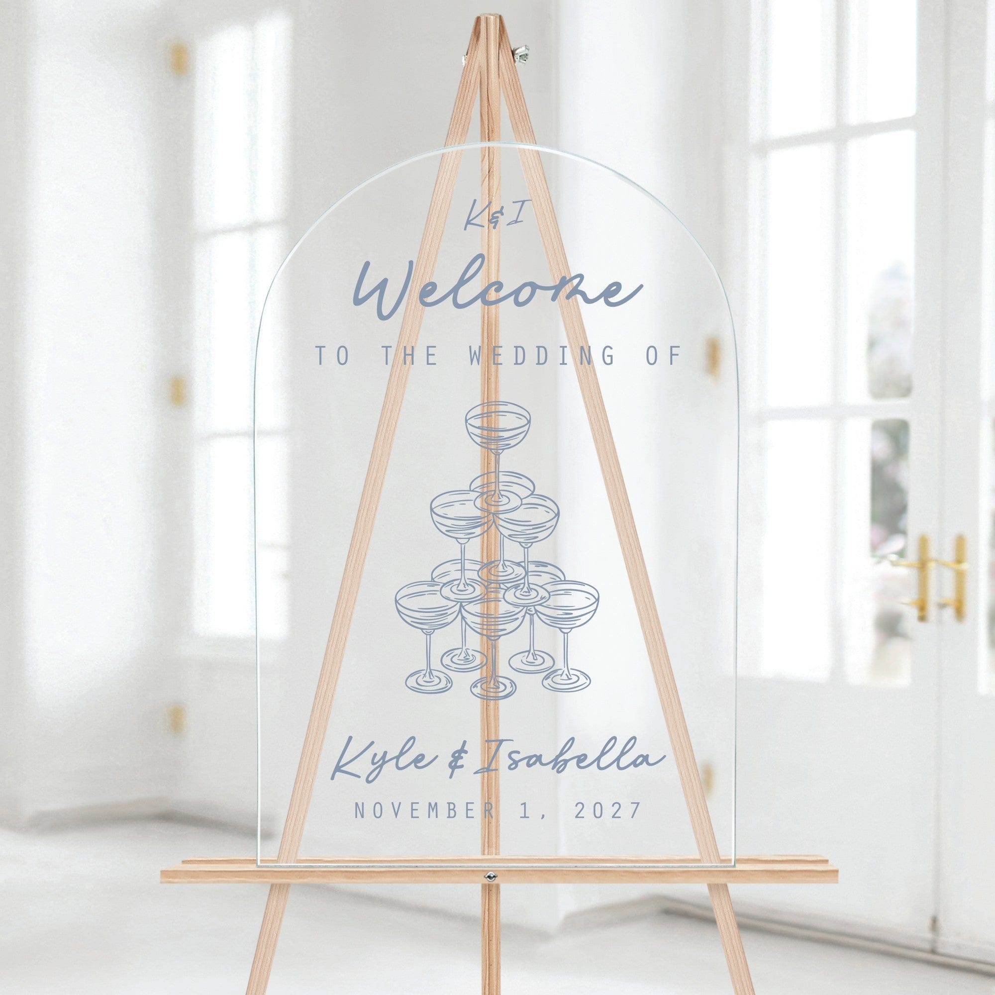 An arched clear acrylic wedding welcome sign featuring a light blue champagne glass tower graphic and custom welcome text, displayed on a light wood easel.