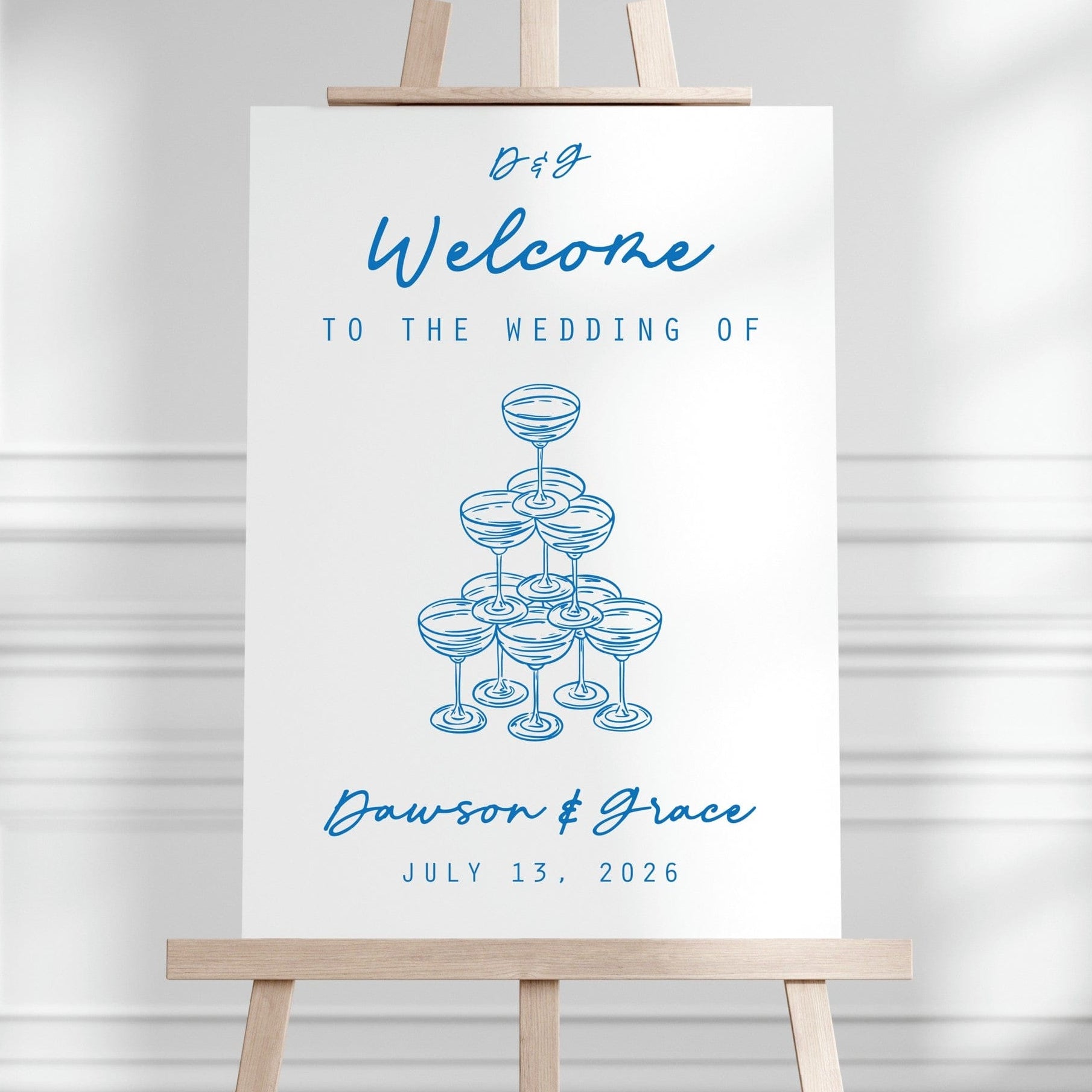 A white wedding welcome sign displays blue script text and a blue line art illustration of stacked champagne coupes, presented on a light brown wooden easel.