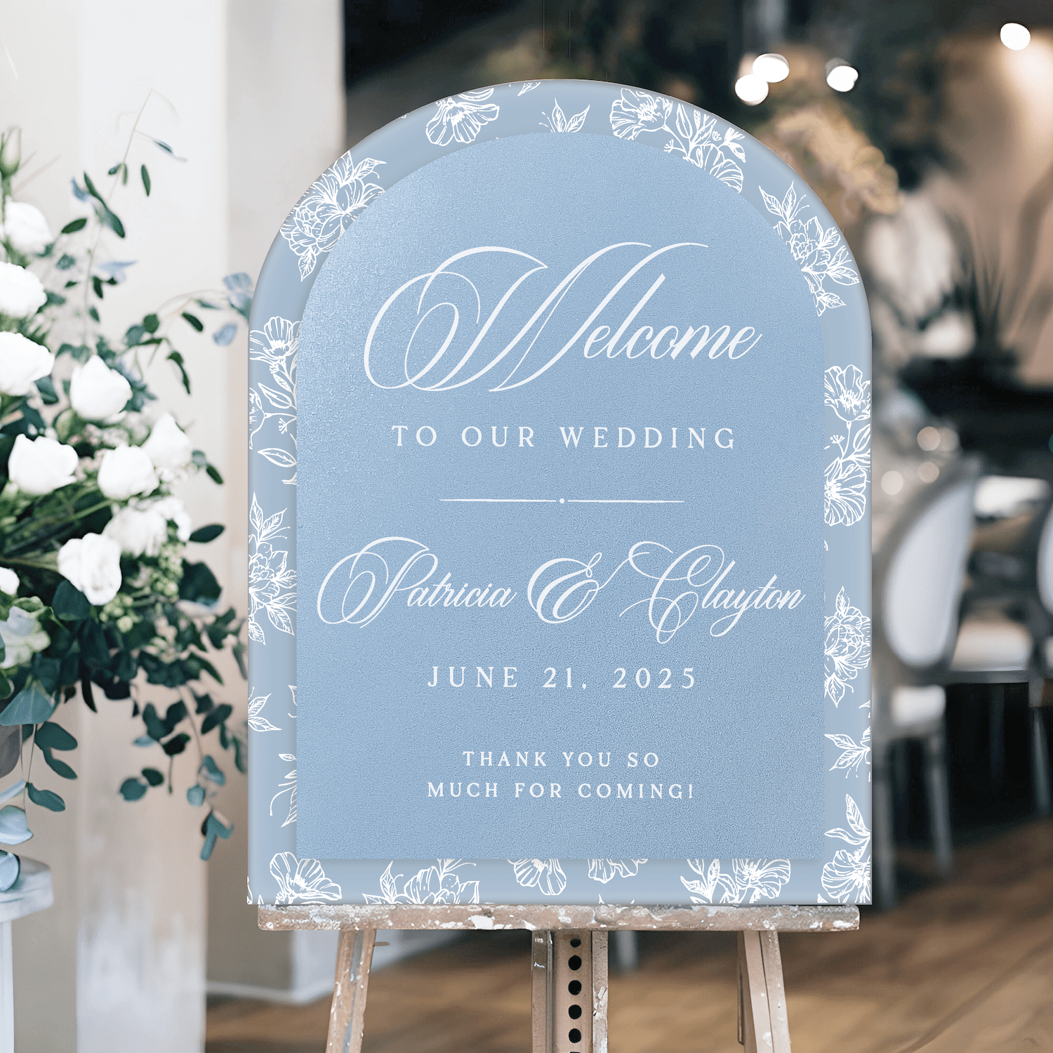Blue arch-shaped wedding welcome sign with white toile floral border and script text reading "Welcome to Our Wedding" with couple's names and date June 21, 2025.