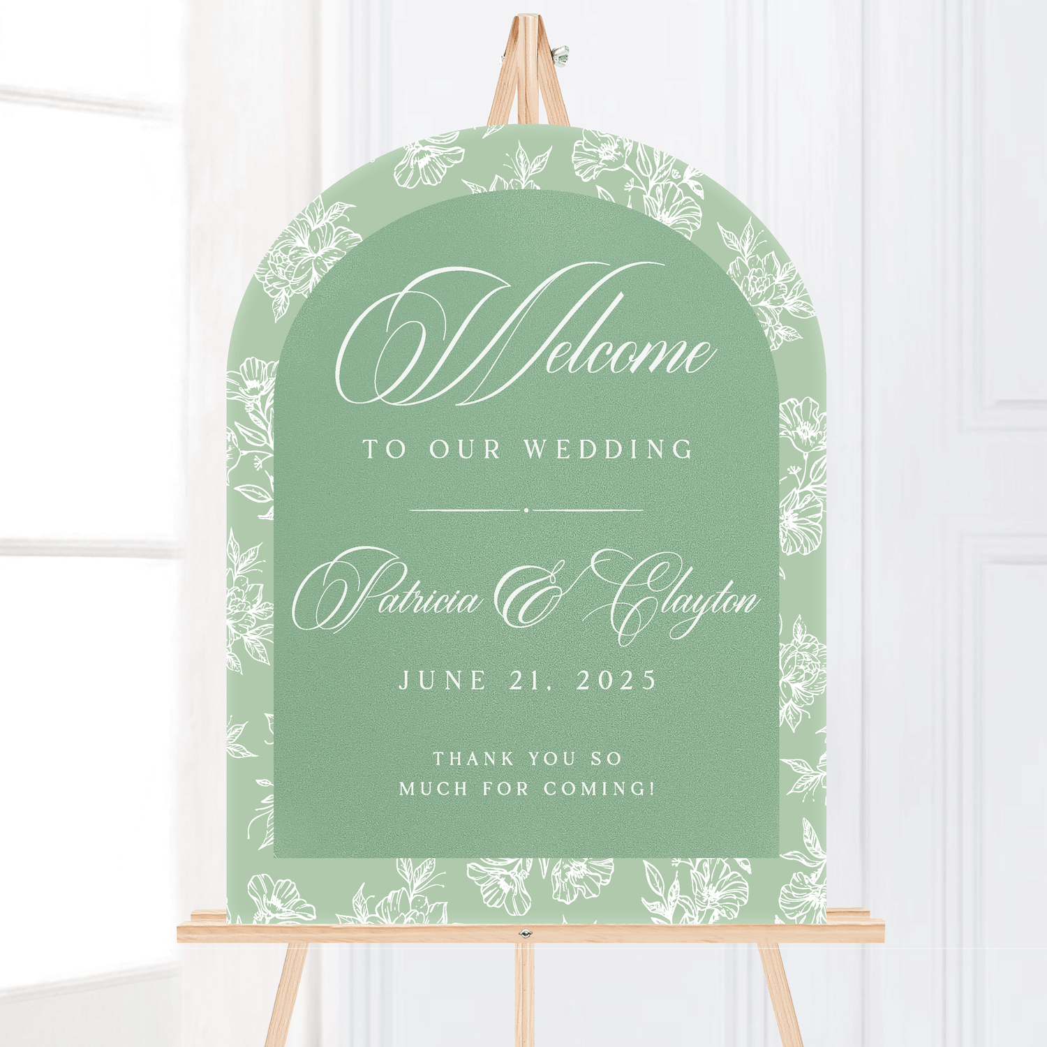 Sage green arch-shaped welcome sign with white toile floral border, personalized with couple names and wedding date, displayed on wooden easel.