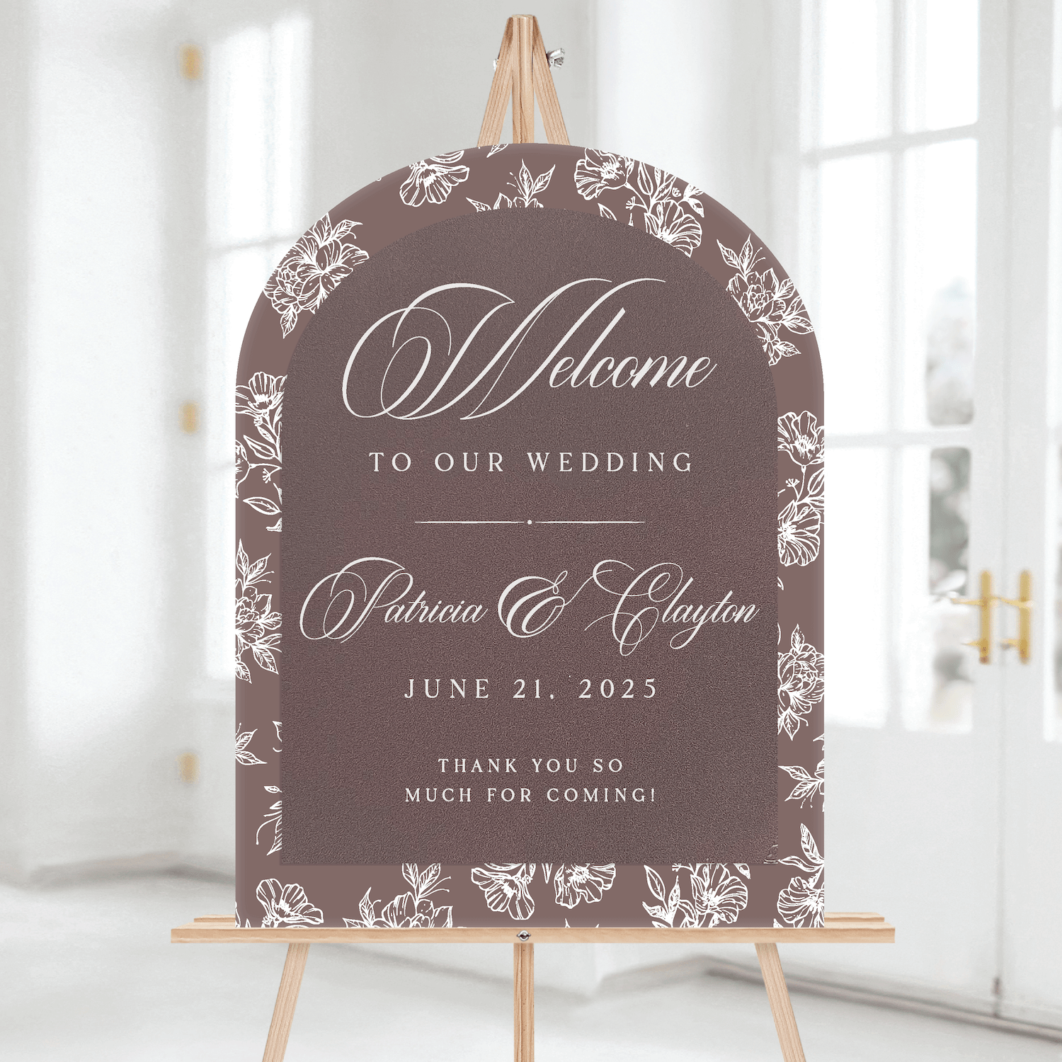 Arched gray wedding welcome sign with white floral toile border and elegant script reading "Welcome to our wedding" with couple's names and June 21, 2025 date, displayed on wooden easel.