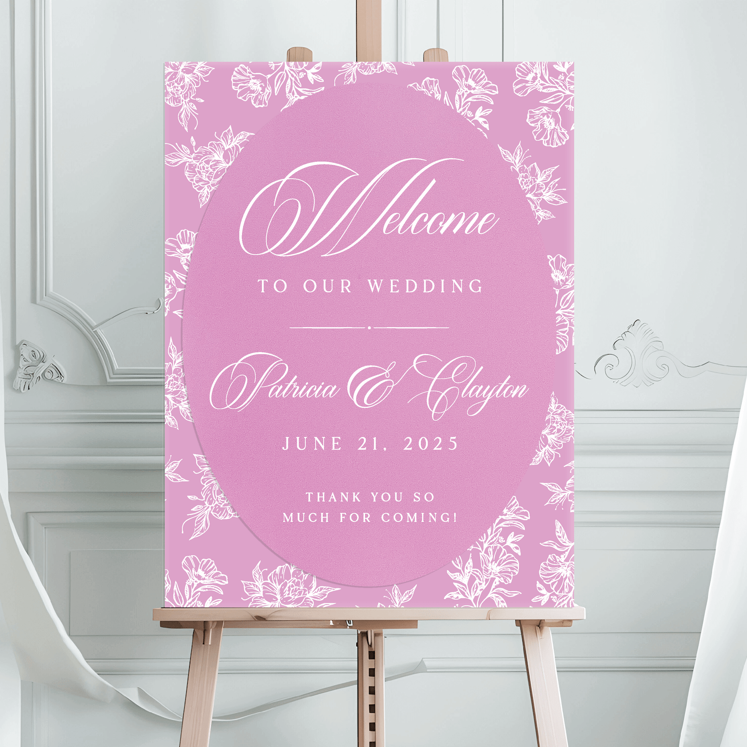 Pink floral wedding welcome sign with white toile pattern border, script text reading "Welcome to our wedding" and couple names "Adriana & Clayton" with date "June 21, 2025" on wooden easel.
