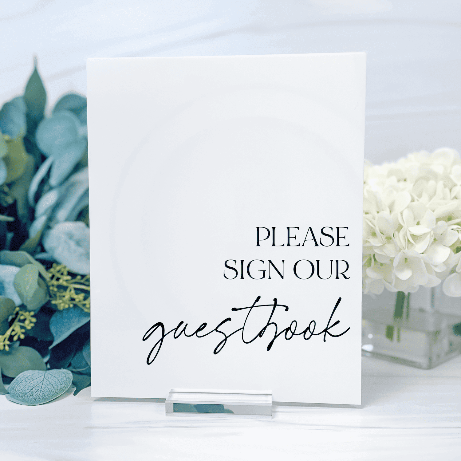 White acrylic table sign with "Please sign our guestbook" text in black and script font, displayed with eucalyptus and white flowers.