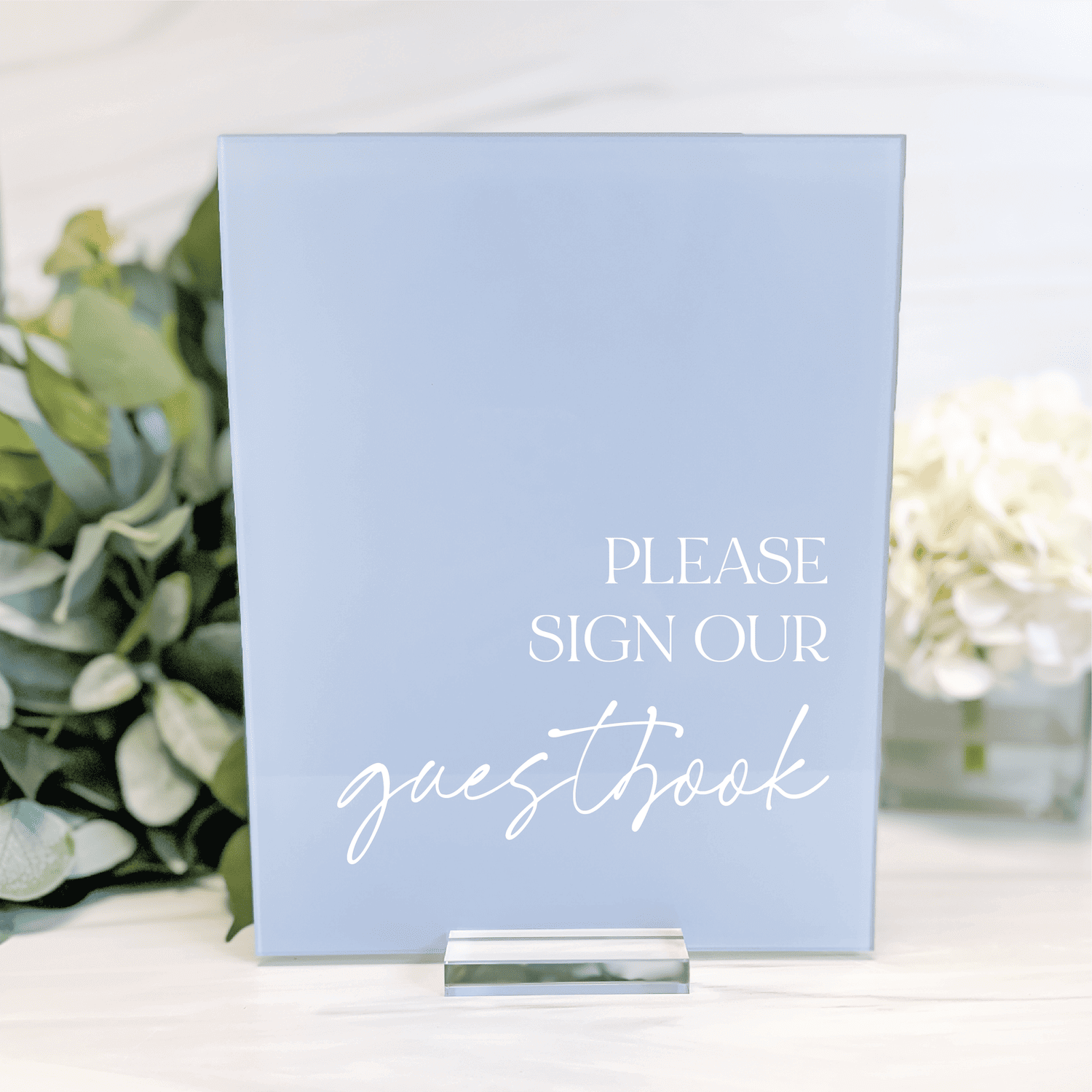 Light blue acrylic table sign with white text reading "Please Sign Our Guestbook" displayed next to flowers and greenery