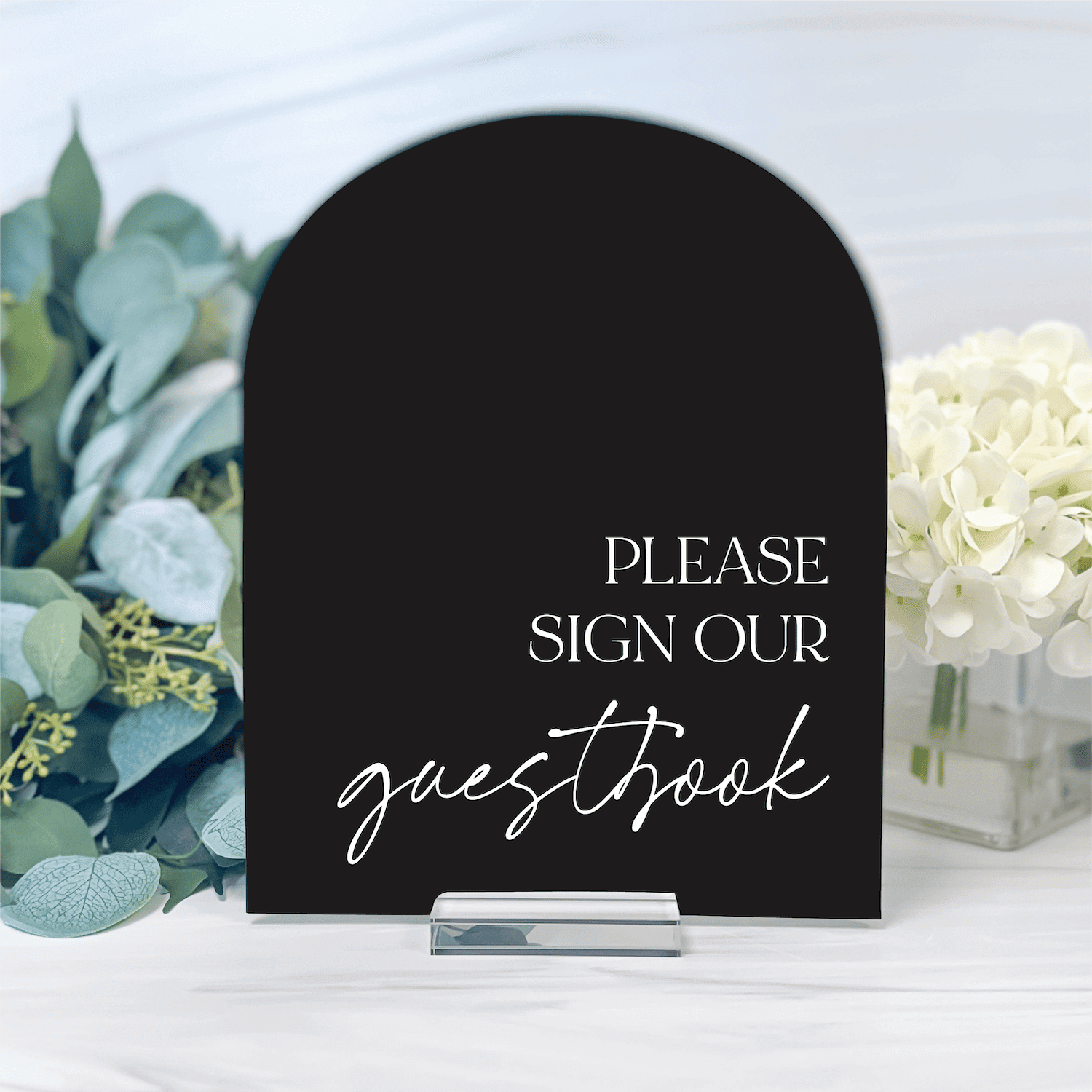 Black acrylic arch-shaped table sign with "Please sign our guestbook" text in white, displayed with eucalyptus and white flowers.