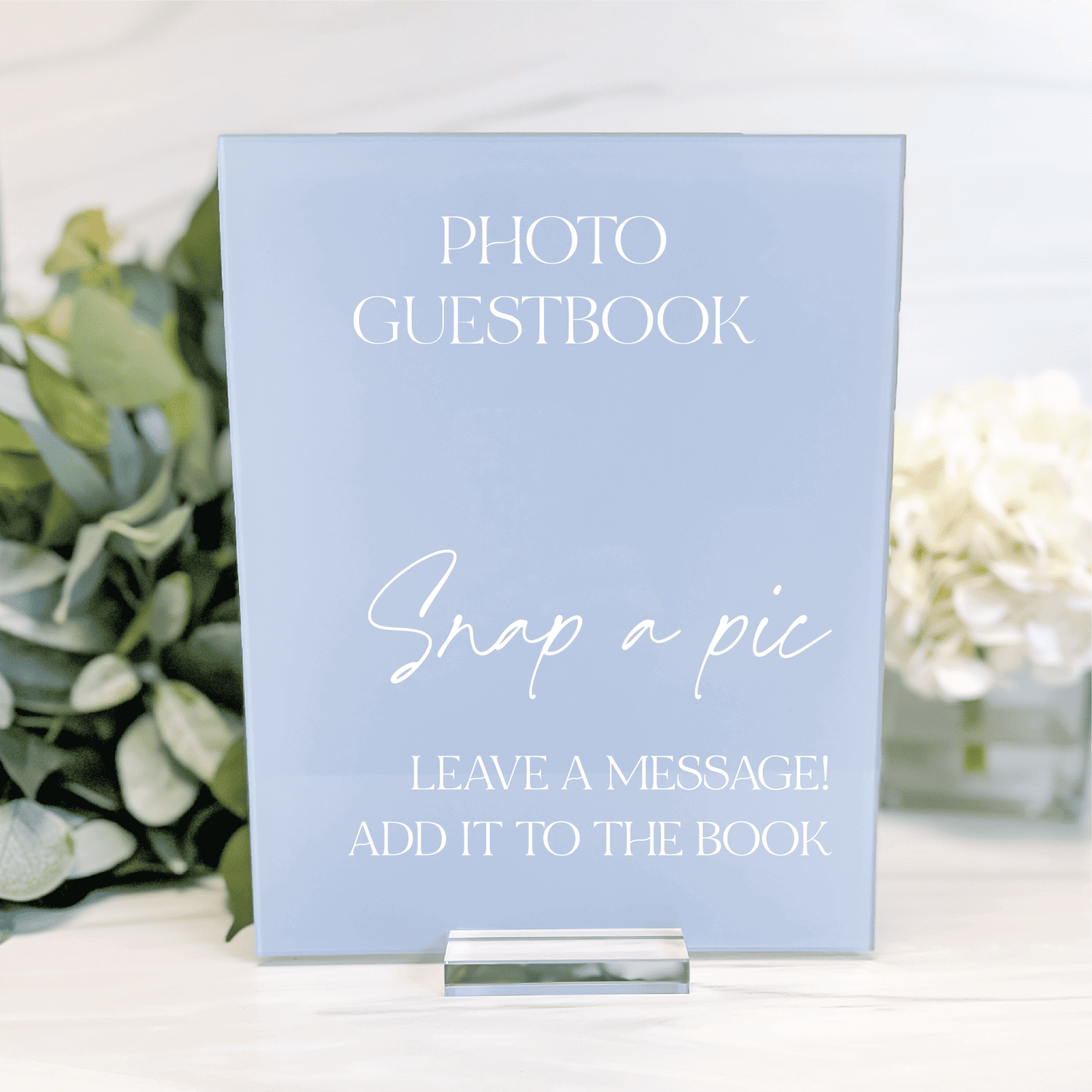 Light blue acrylic table sign with white text reading "Photo Guestbook" and "Snap a pic Leave a message Add to the book" displayed on stand.