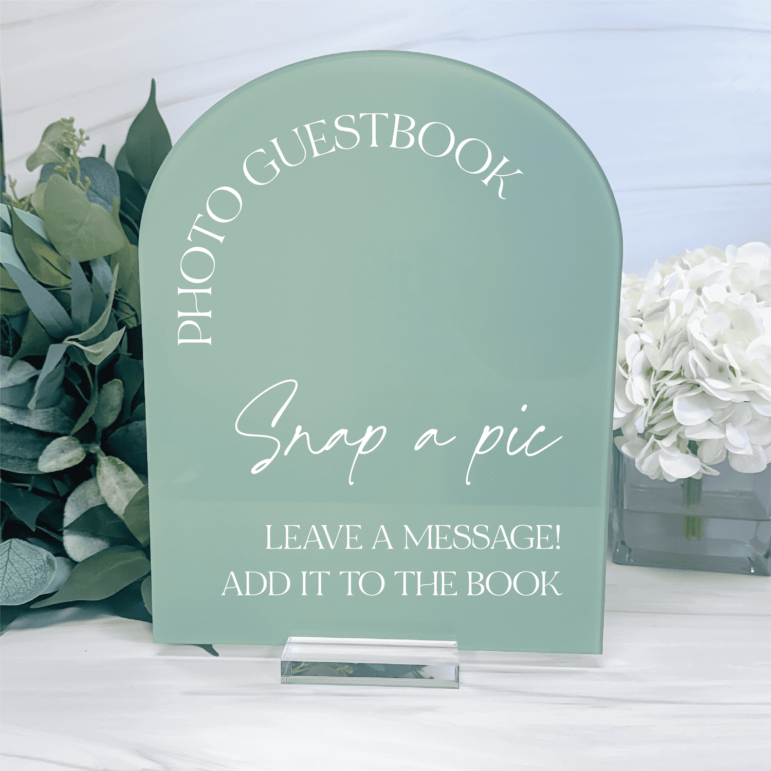 Sage green arched acrylic sign with white text reading "Photo Guestbook," "Snap a pic," and "Leave a message! Add it to the book," surrounded by eucalyptus and white flowers.