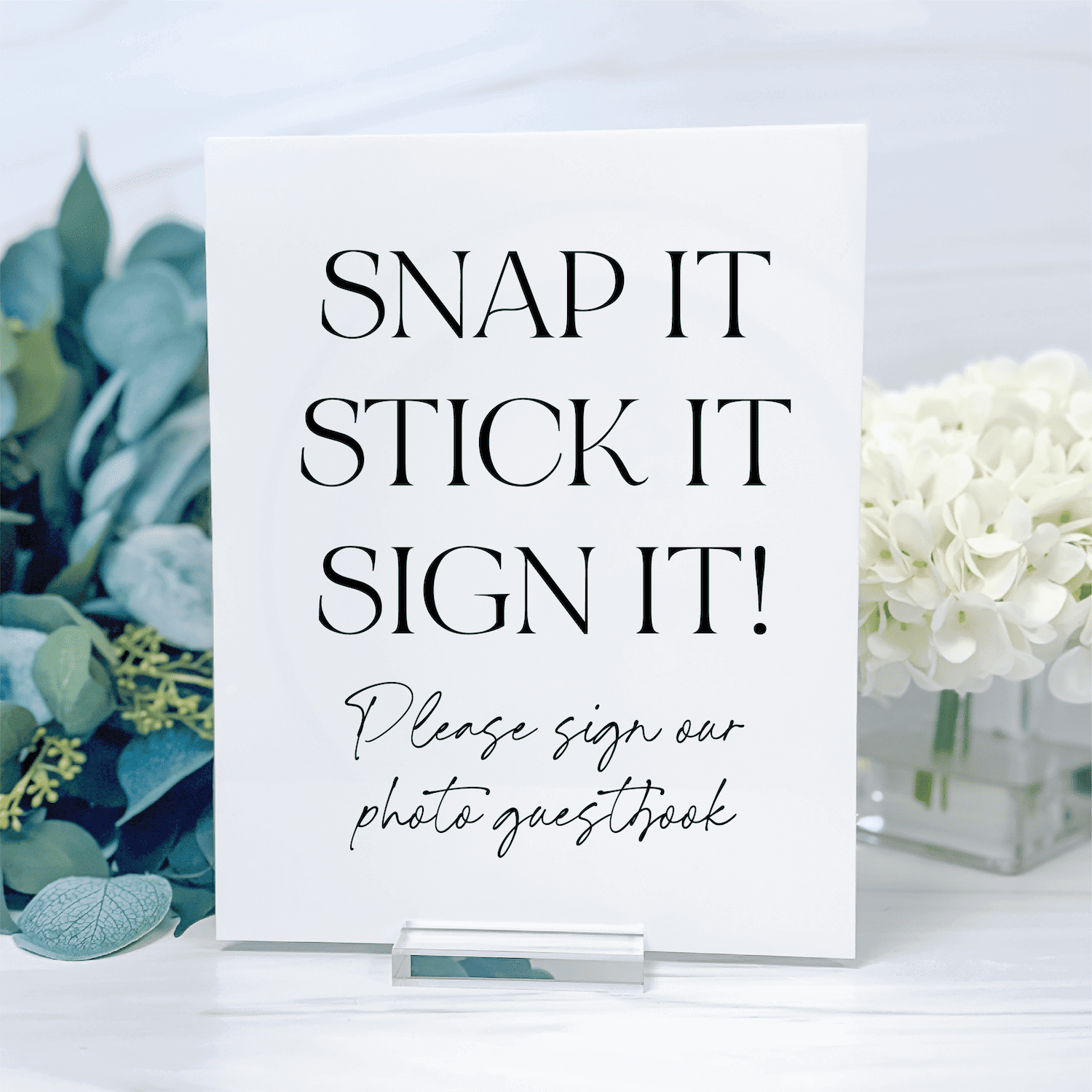White acrylic sign with "Snap It Stick It Sign It" text and photo guestbook instructions, displayed with eucalyptus and white flowers.
