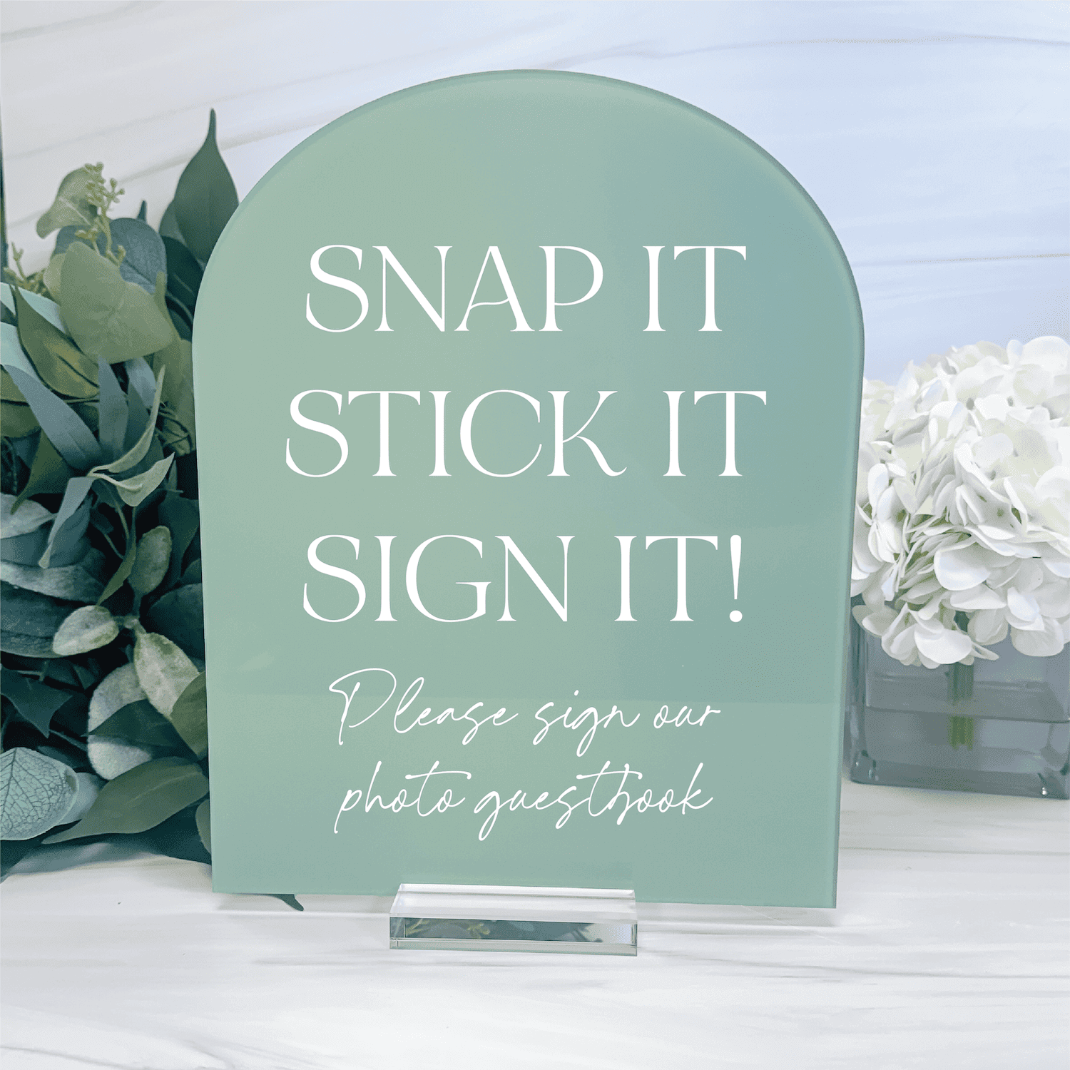 Sage green acrylic arch sign with white text reading "Snap it Stick it Sign it" and "Please sign our photo guestbook" displayed on white table with flowers.