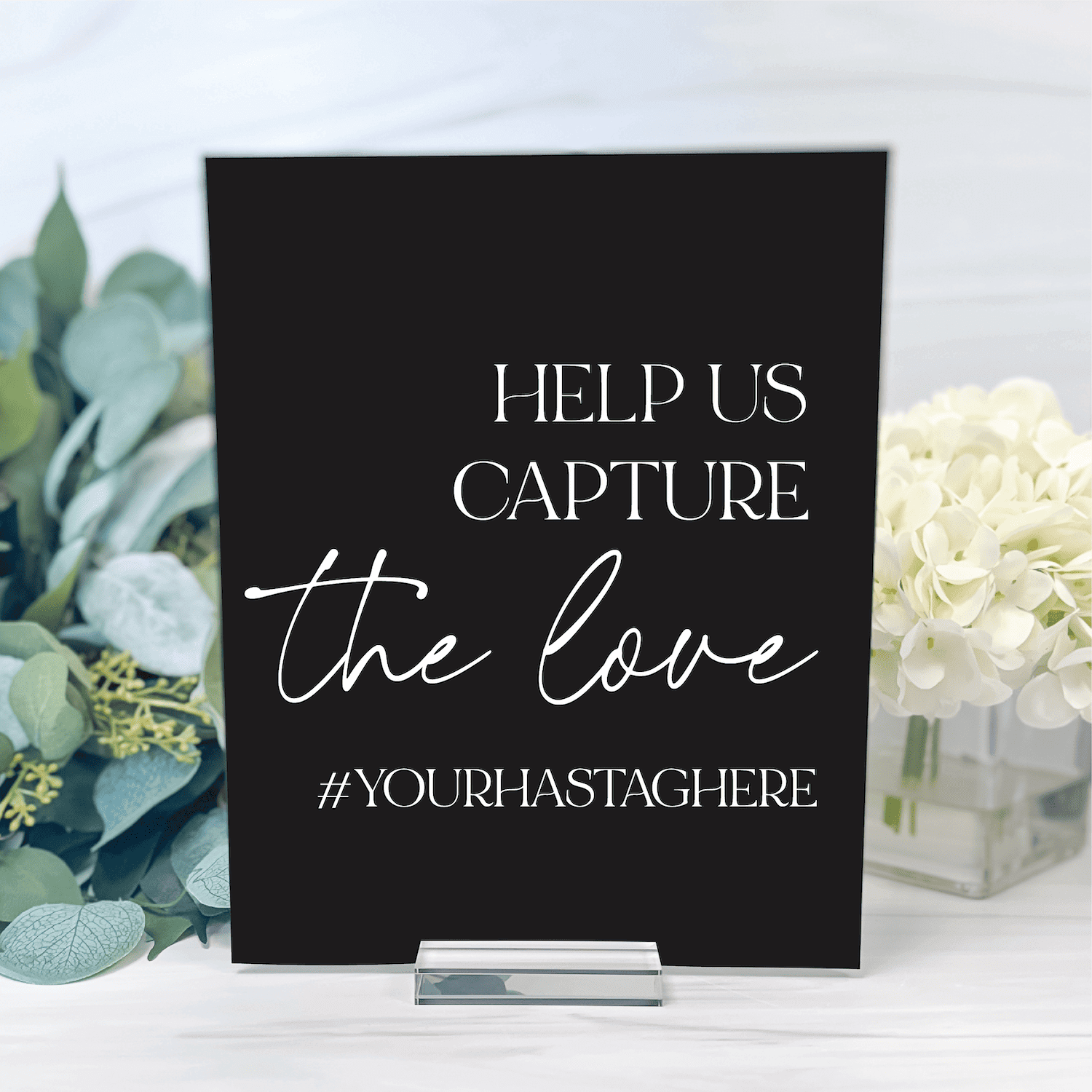 Black acrylic table sign with white text reading "Help us capture the love" and hashtag placeholder, displayed between white flowers and greenery.