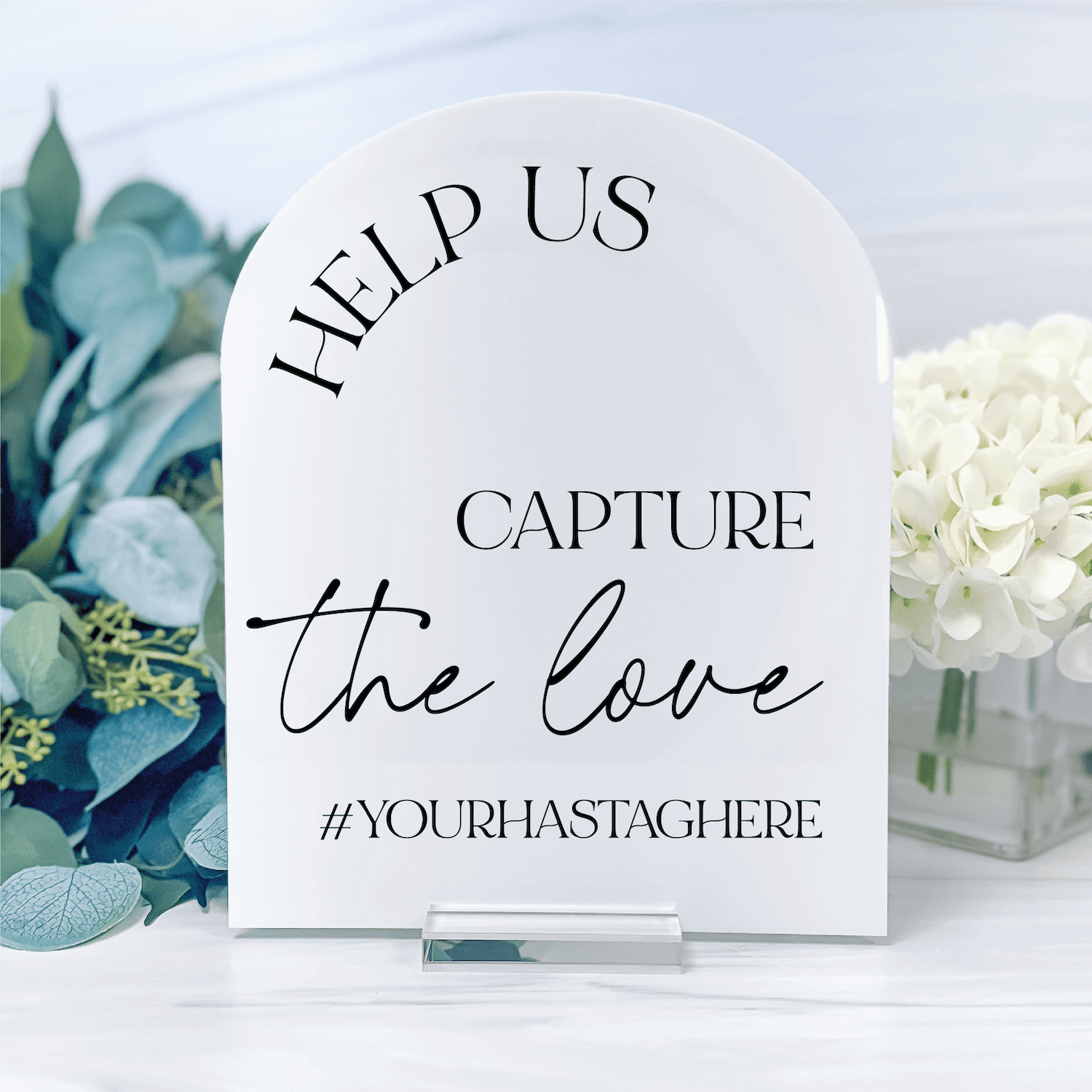 White arched acrylic sign reading "Help us capture the love #yourhastaghere" with blue eucalyptus and white flowers beside it