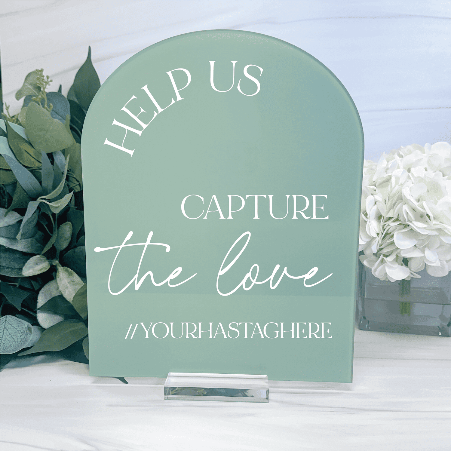 Sage green arch-shaped acrylic sign with white text reading "Help us capture the love #yourhastagherehere" surrounded by eucalyptus and white flowers.