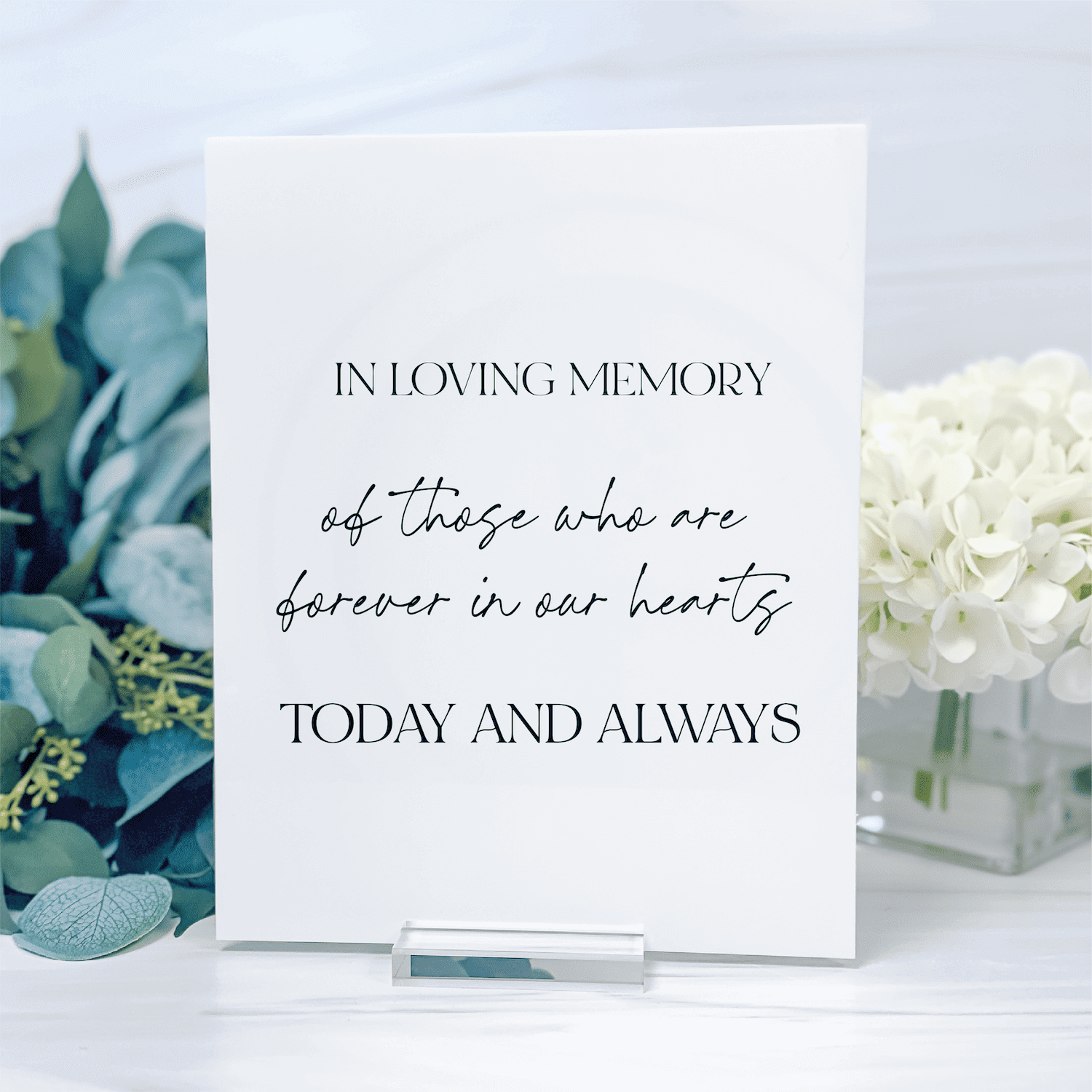 White acrylic memorial sign with "In Loving Memory" text and script typography, displayed on a table with eucalyptus and white flowers.