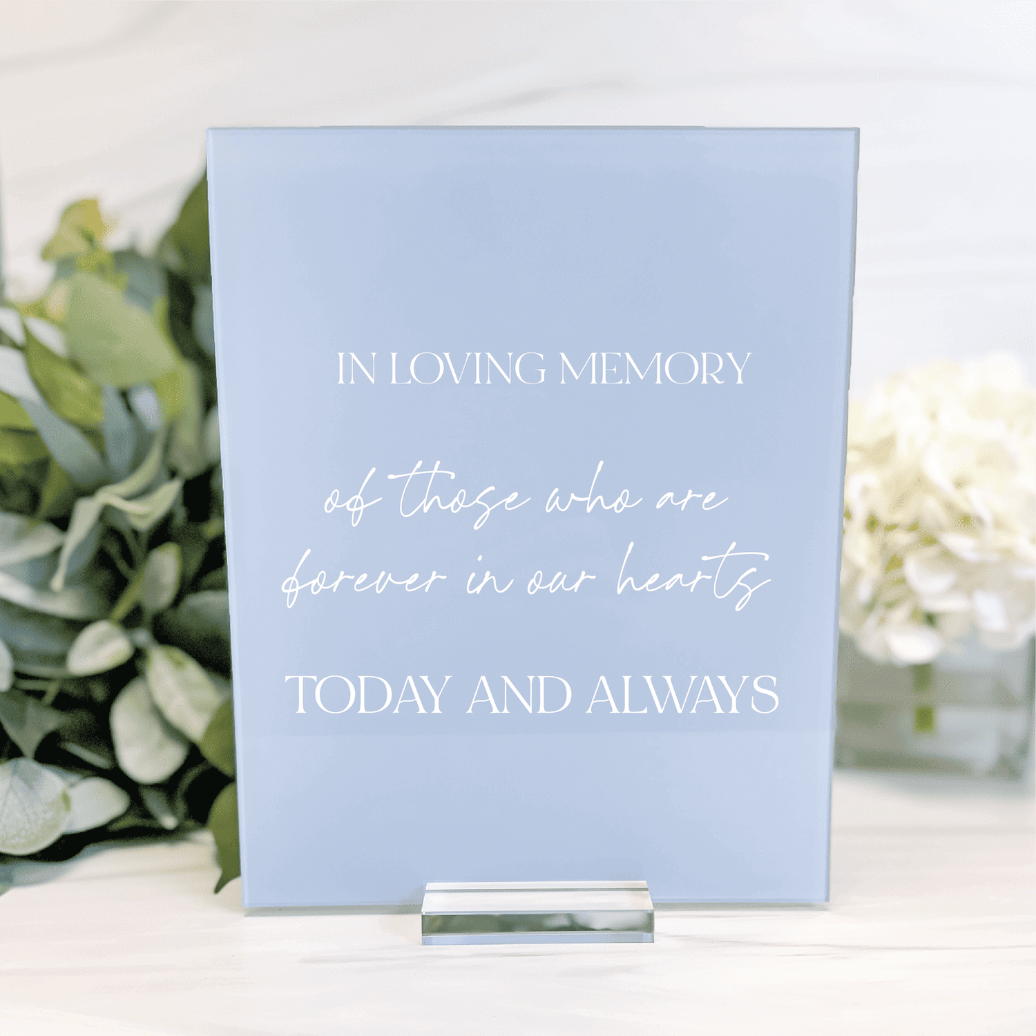 Light blue acrylic memorial sign reading "In Loving Memory of those who are forever in our hearts Today and Always" with silver stand, displayed with white flowers