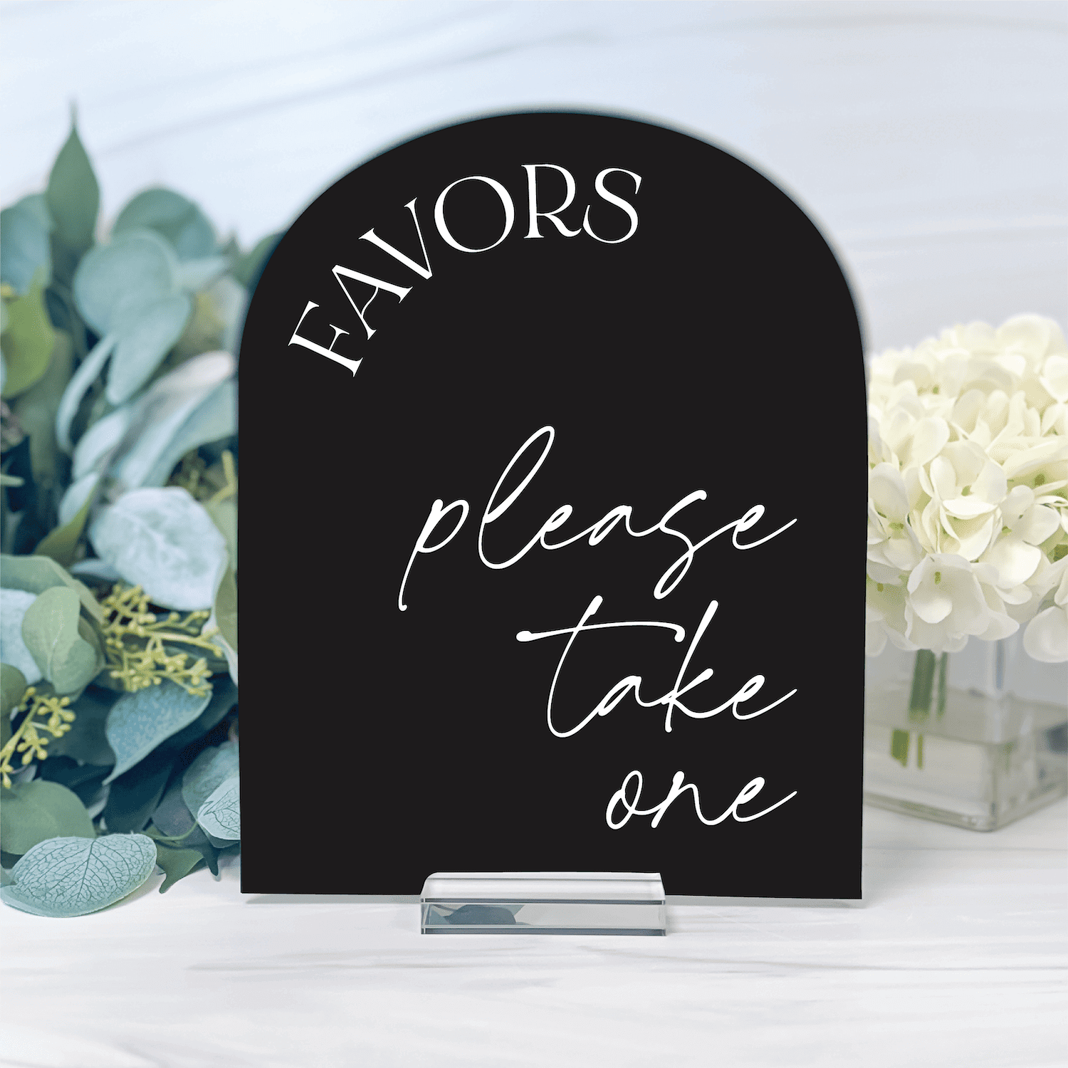 Black arch-shaped acrylic sign with white script text reading "FAVORS please take one" displayed with flowers and greenery.