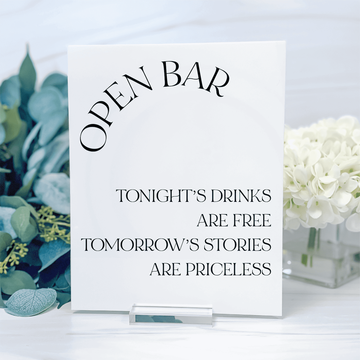 White acrylic sign with "OPEN BAR" text and quote "Tonight's drinks are free, tomorrow's stories are priceless" displayed on a table with eucalyptus and white flowers.