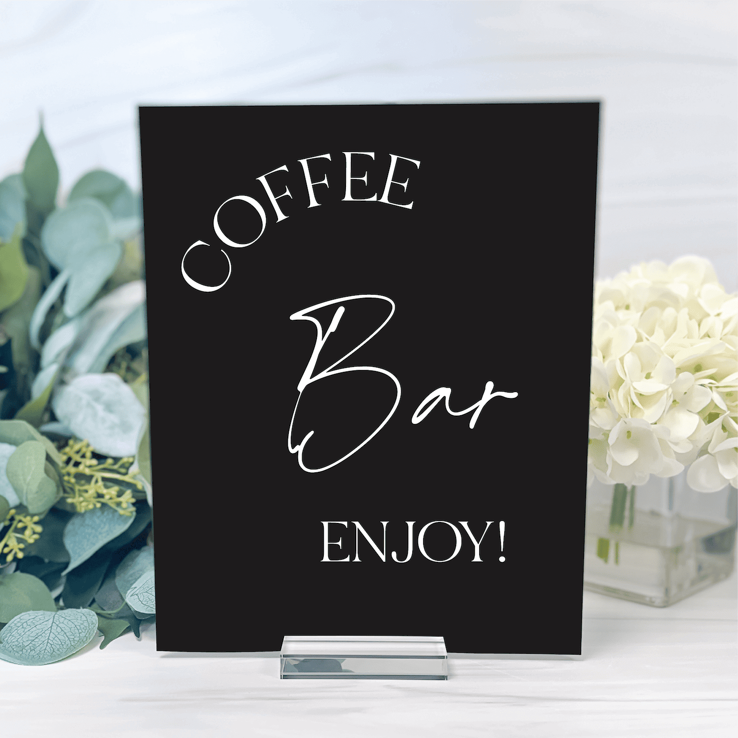 Black acrylic table sign with white script text reading "Coffee Bar Enjoy!" displayed with eucalyptus and white flowers.