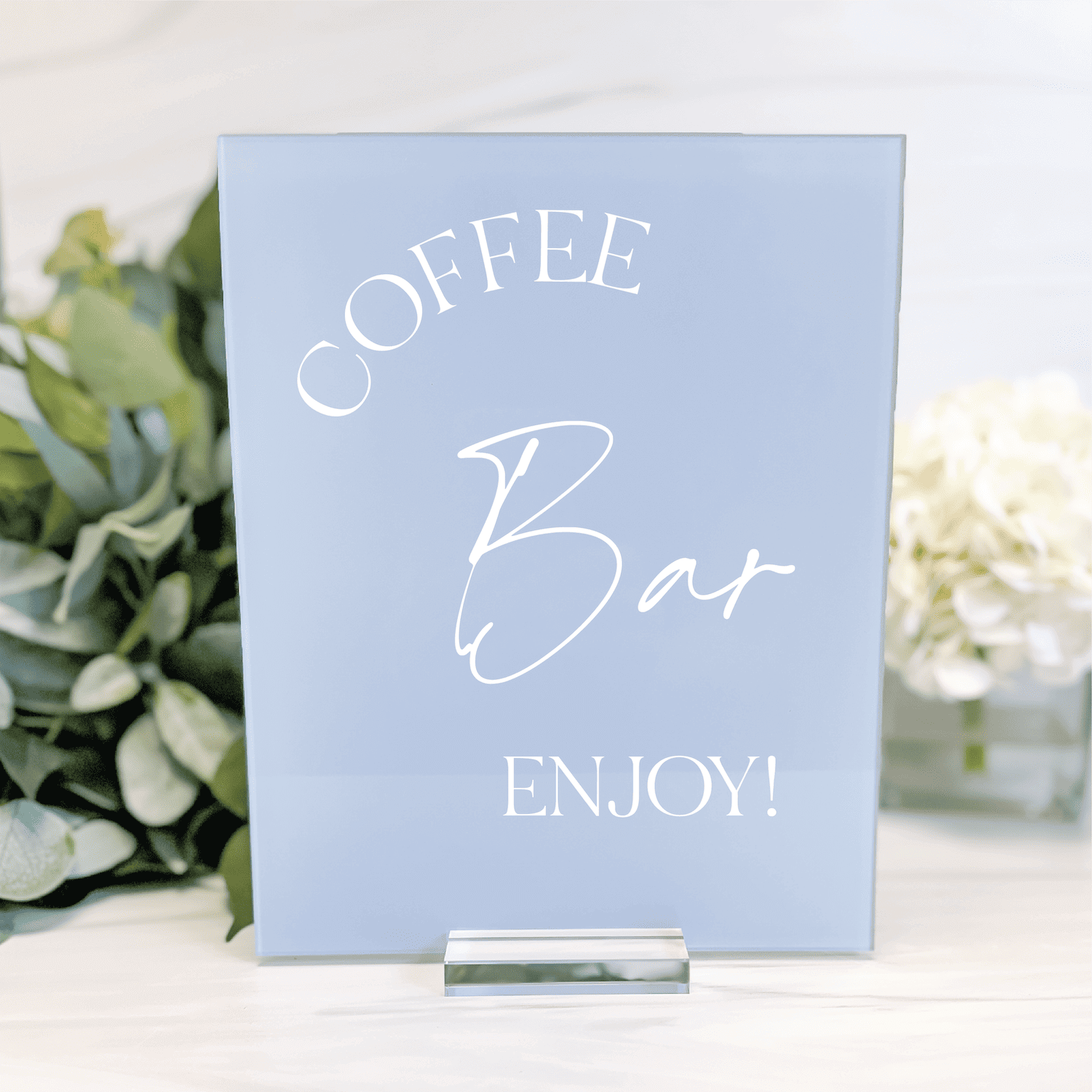 Light blue acrylic table sign with white text reading "Coffee Bar Enjoy!" next to green potted plant and white flowers.