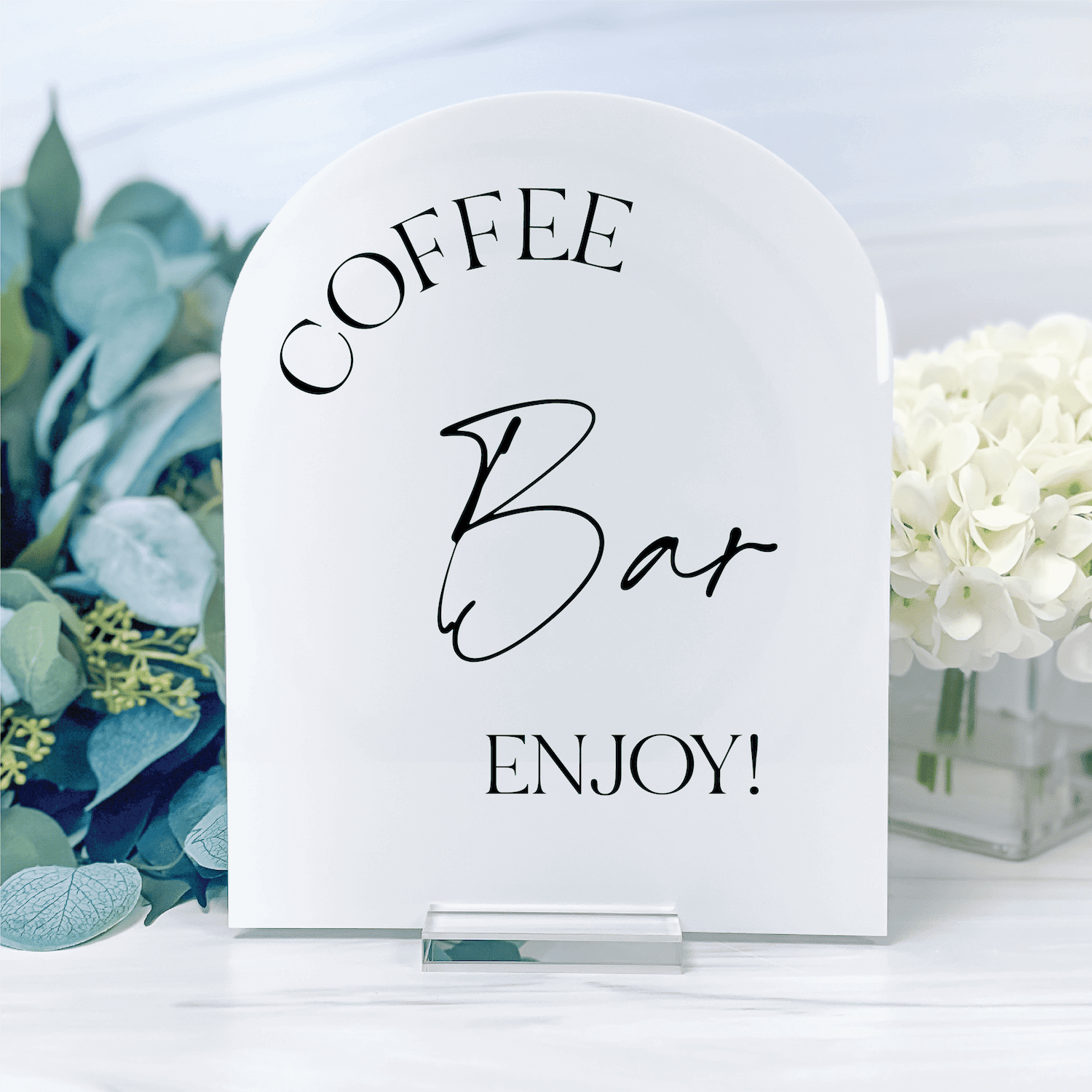 White arched acrylic table sign with "Coffee Bar Enjoy!" text in black lettering, displayed with eucalyptus and white flowers.