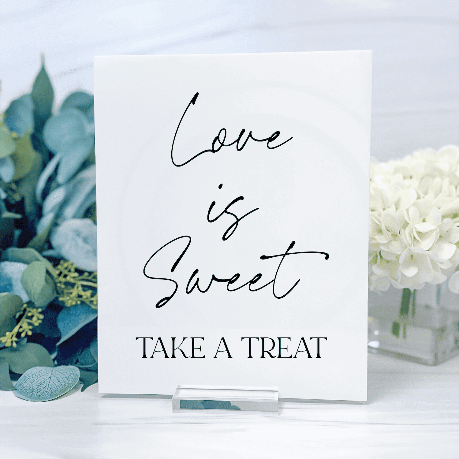 White acrylic sign with "Love is Sweet TAKE A TREAT" in black script and print text, displayed with eucalyptus and white flowers.