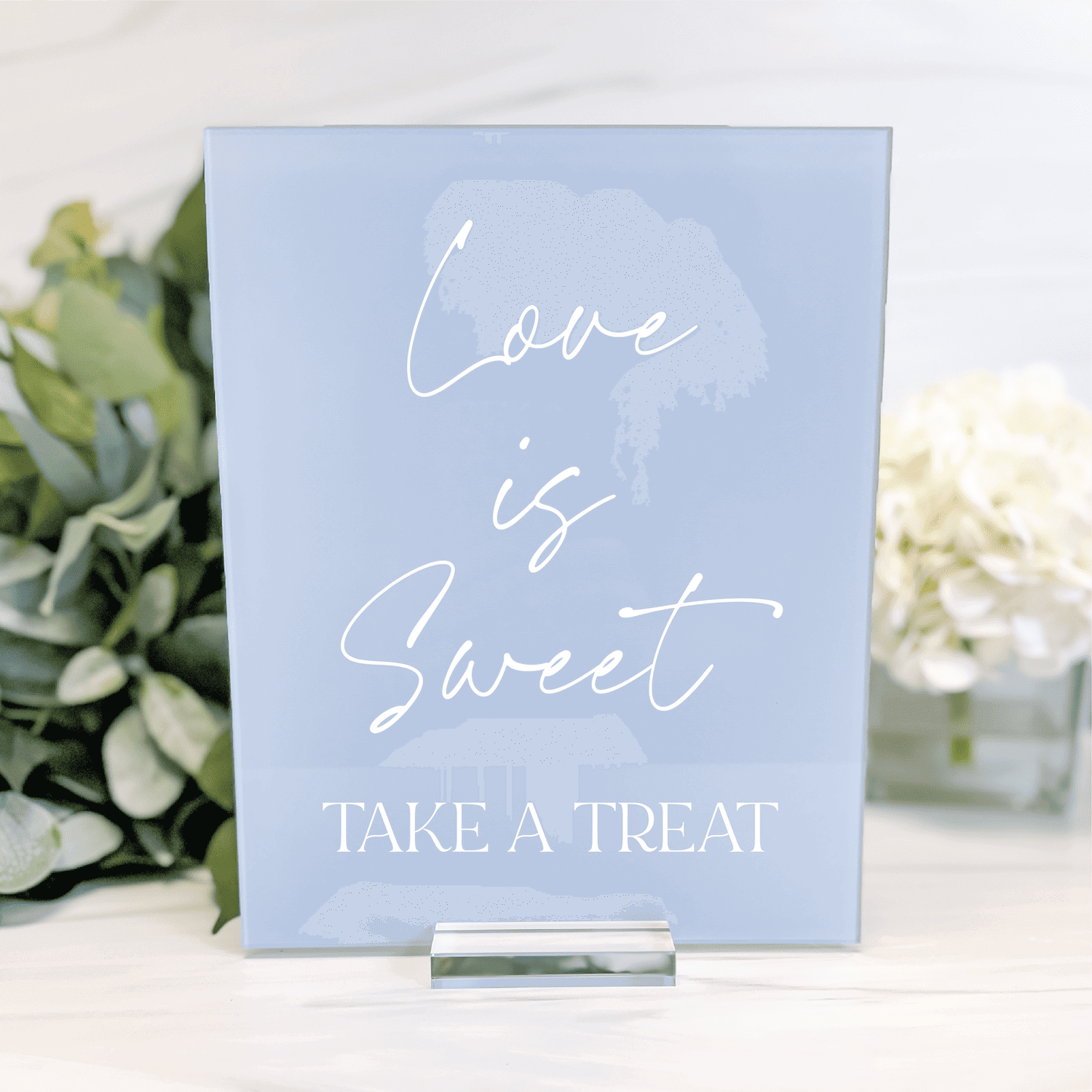 Light blue acrylic sign with white script text reading "Love is Sweet" and "Take a Treat" displayed on a stand with greenery and flowers in background.