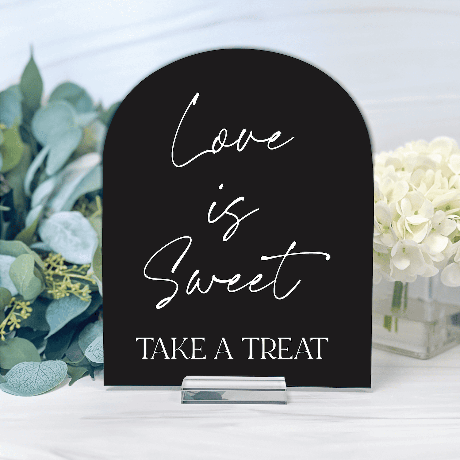 Black arched acrylic sign with "Love is Sweet Take a Treat" text, displayed with greenery and white flowers.