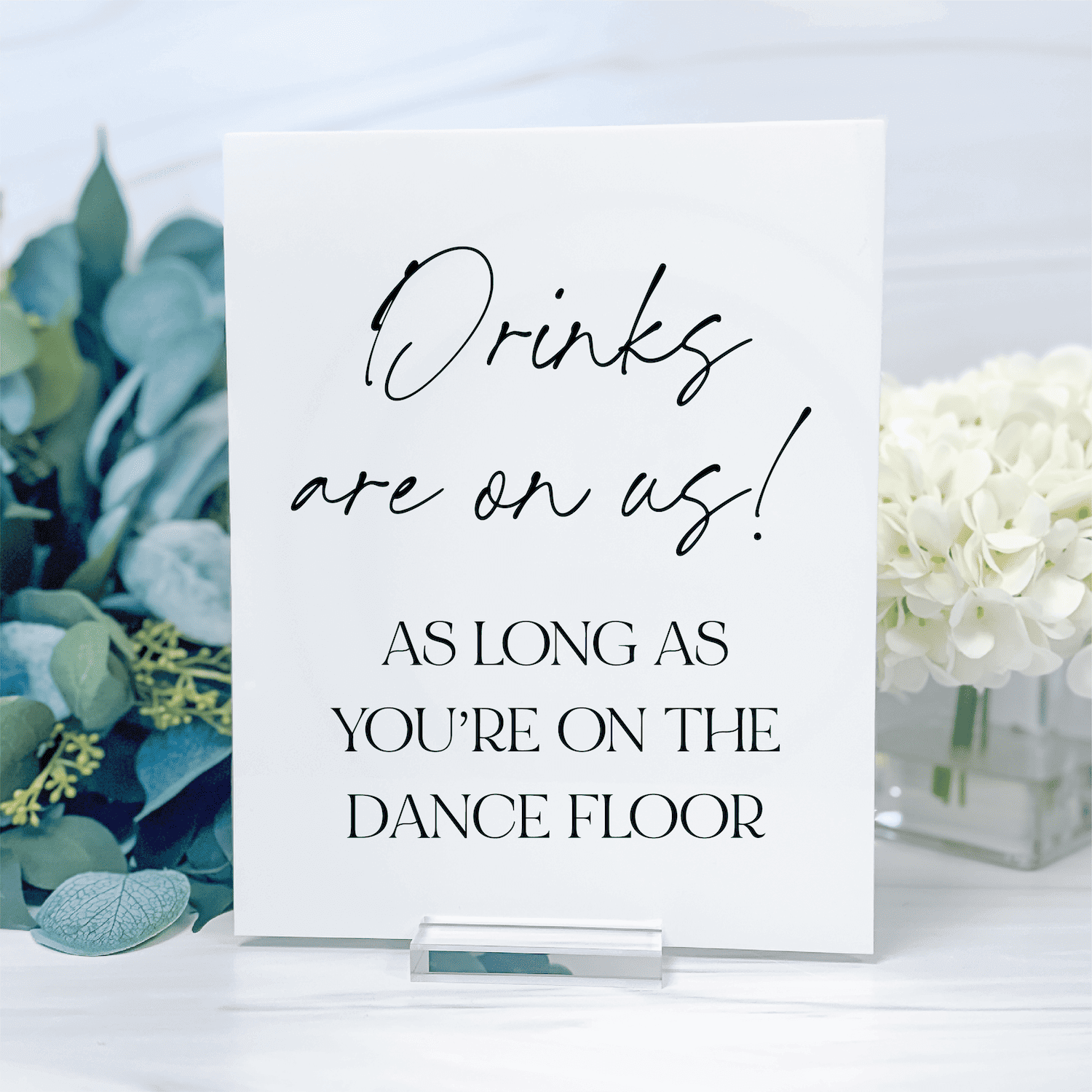 White acrylic table sign with black script and block lettering reading "Drinks are on us! As long as you're on the dance floor" next to eucalyptus and white flowers.