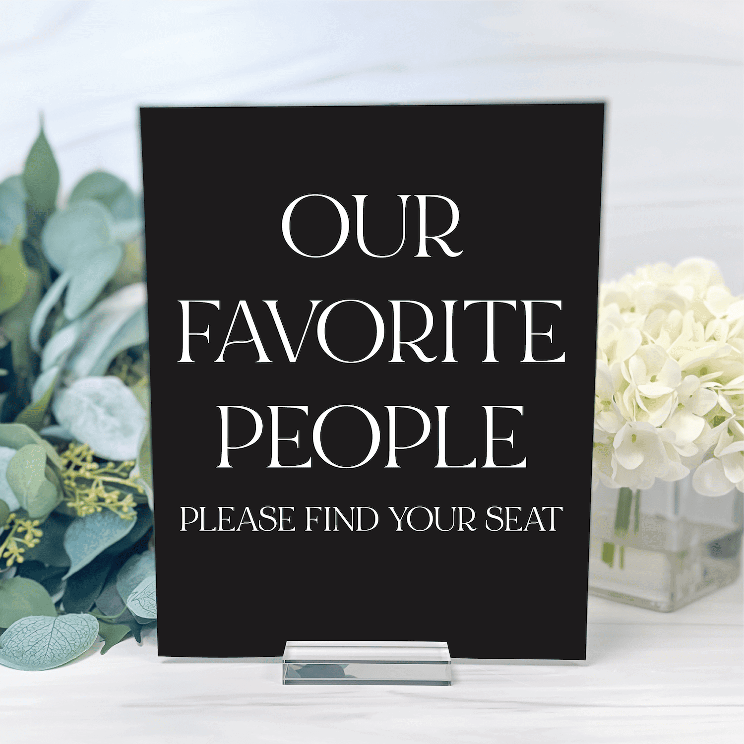 Black acrylic seating sign with white text reading "Our Favorite People Please Find Your Seat" displayed with greenery and white flowers.
