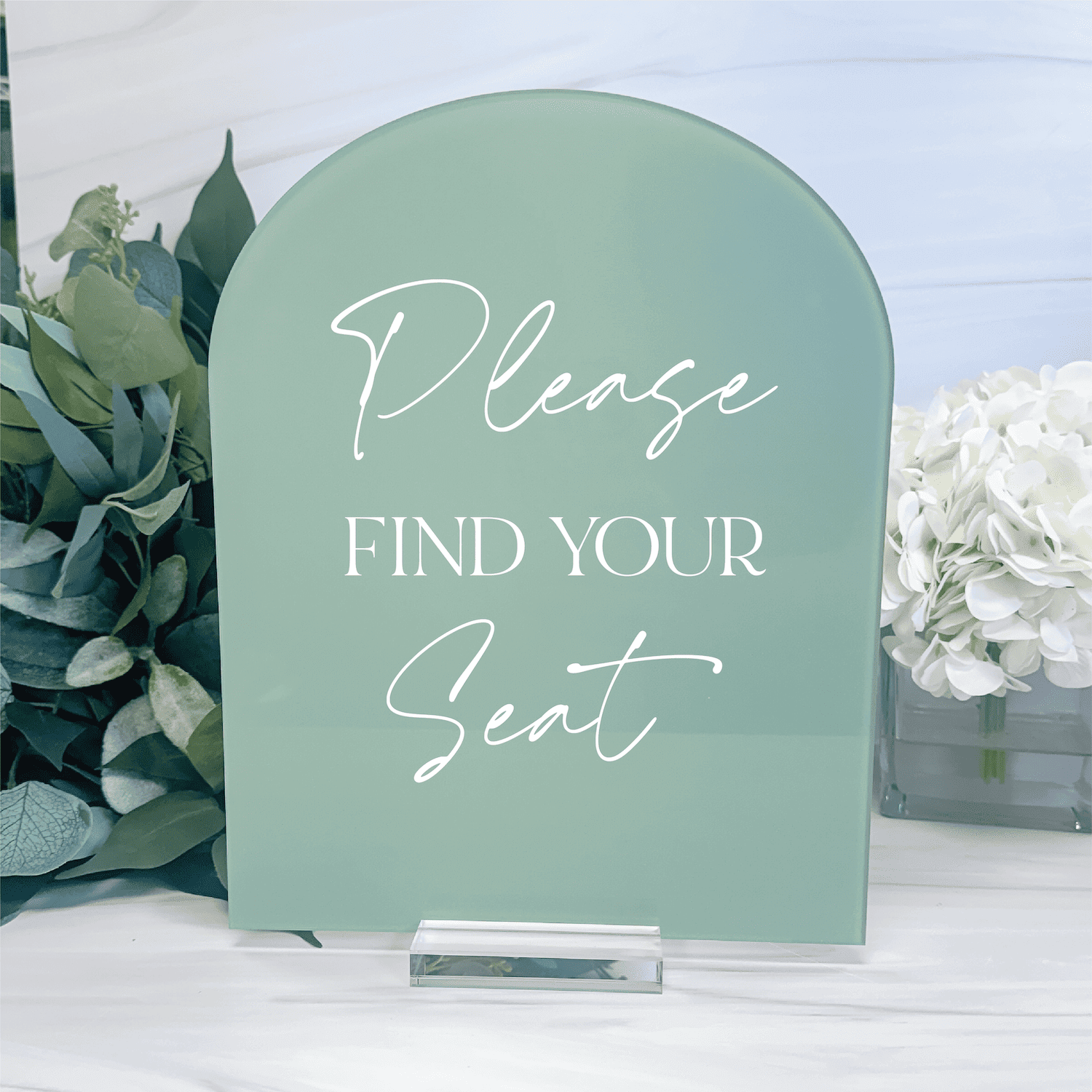 Sage green arched acrylic sign with white script text reading "Please FIND YOUR Seat" displayed with greenery and white flowers.