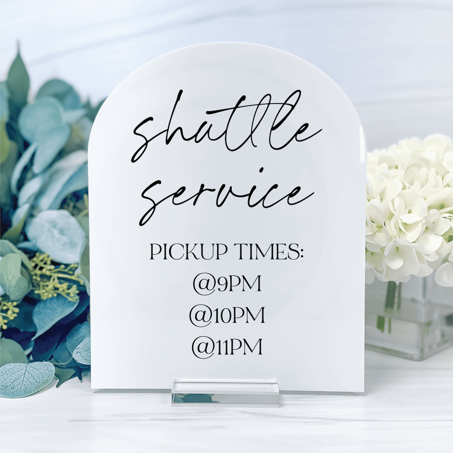White arch-shaped acrylic sign with "shuttle service" in script and pickup times listed below, displayed next to white flowers and green foliage.