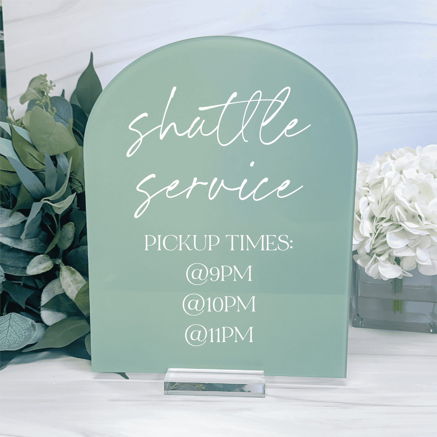 Sage green arched acrylic sign with white script text reading "shuttle service" and pickup times, flanked by eucalyptus and white flowers.