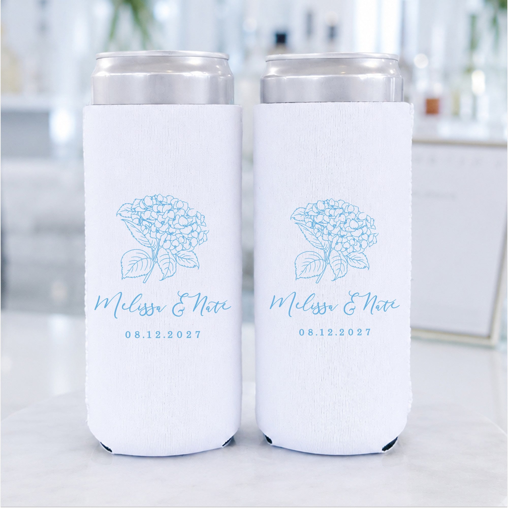 Two white slim can coolers, made of a fabric-like material, feature a delicate light blue hydrangea floral design and are ideal as personalized wedding favors.
