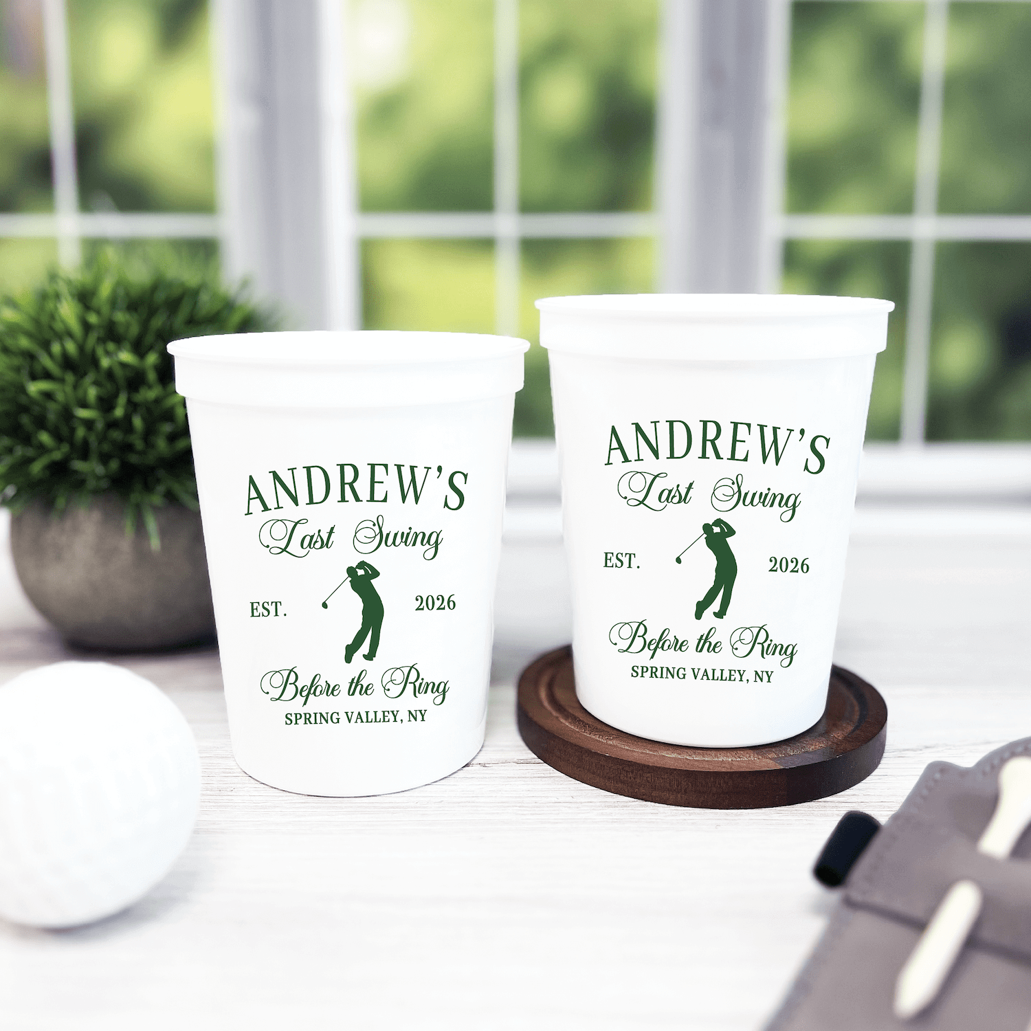 White stadium cups with green golf club design and "Andrew's Last Swing Before The Ring EST. 2026" text, displayed on wooden coasters by a window.