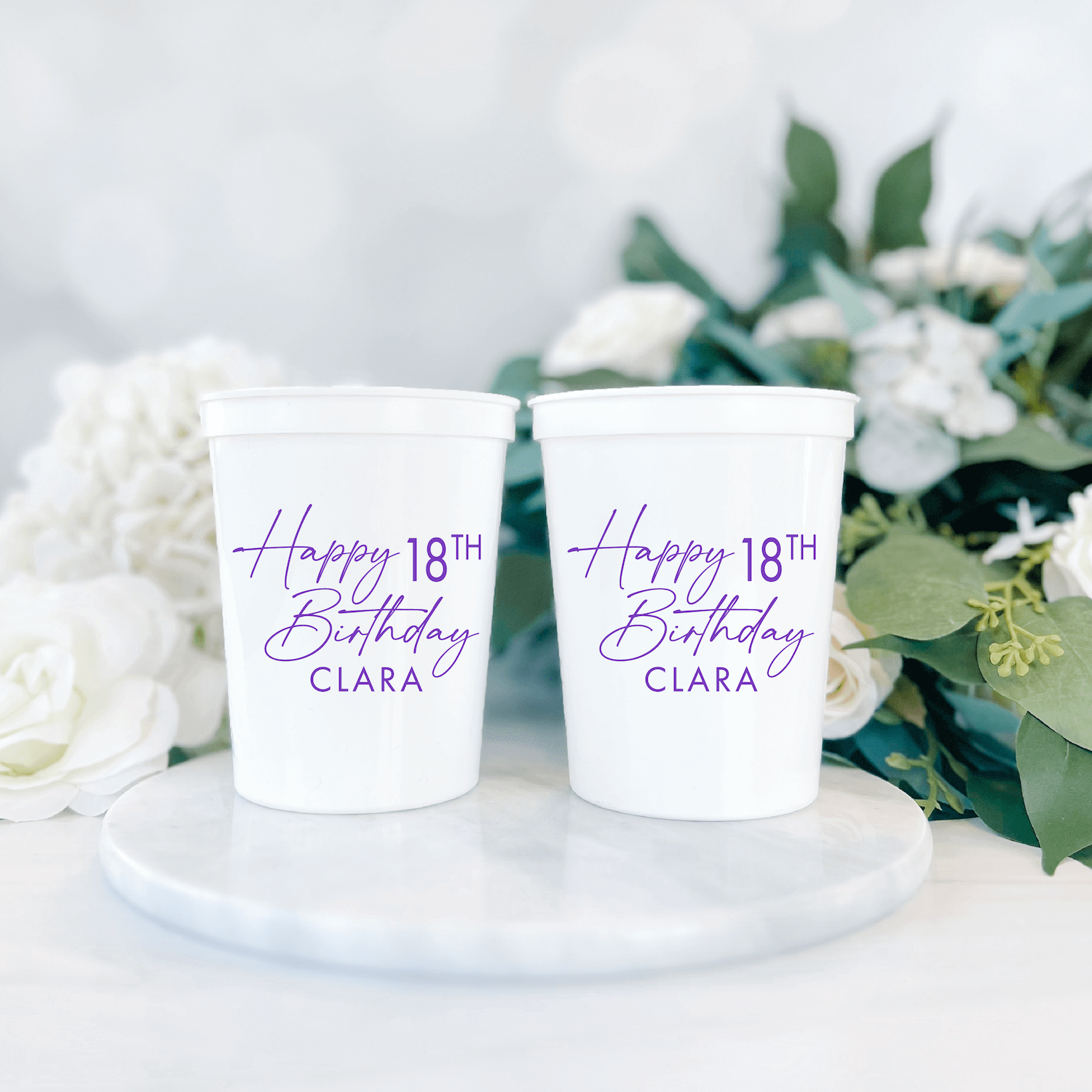 Two white stadium cups with purple script text reading "Happy 18th Birthday Clara" displayed on a marble plate with white flowers.