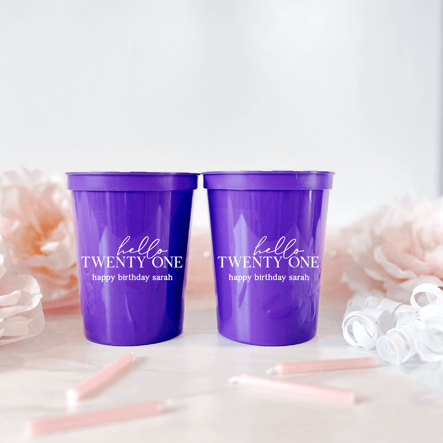 Pair of purple stadium cups with "Hello Twenty-One happy birthday sarah" personalized text in white lettering, displayed with pink flowers and party decorations.