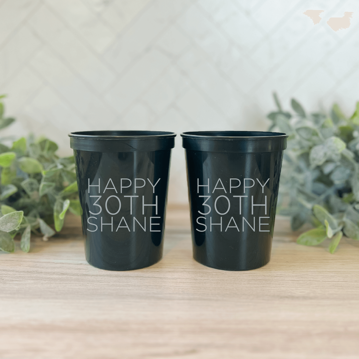 Two black stadium cups personalized with "Happy 30th Shane" in silver lettering, displayed on a wooden surface with greenery in the background.