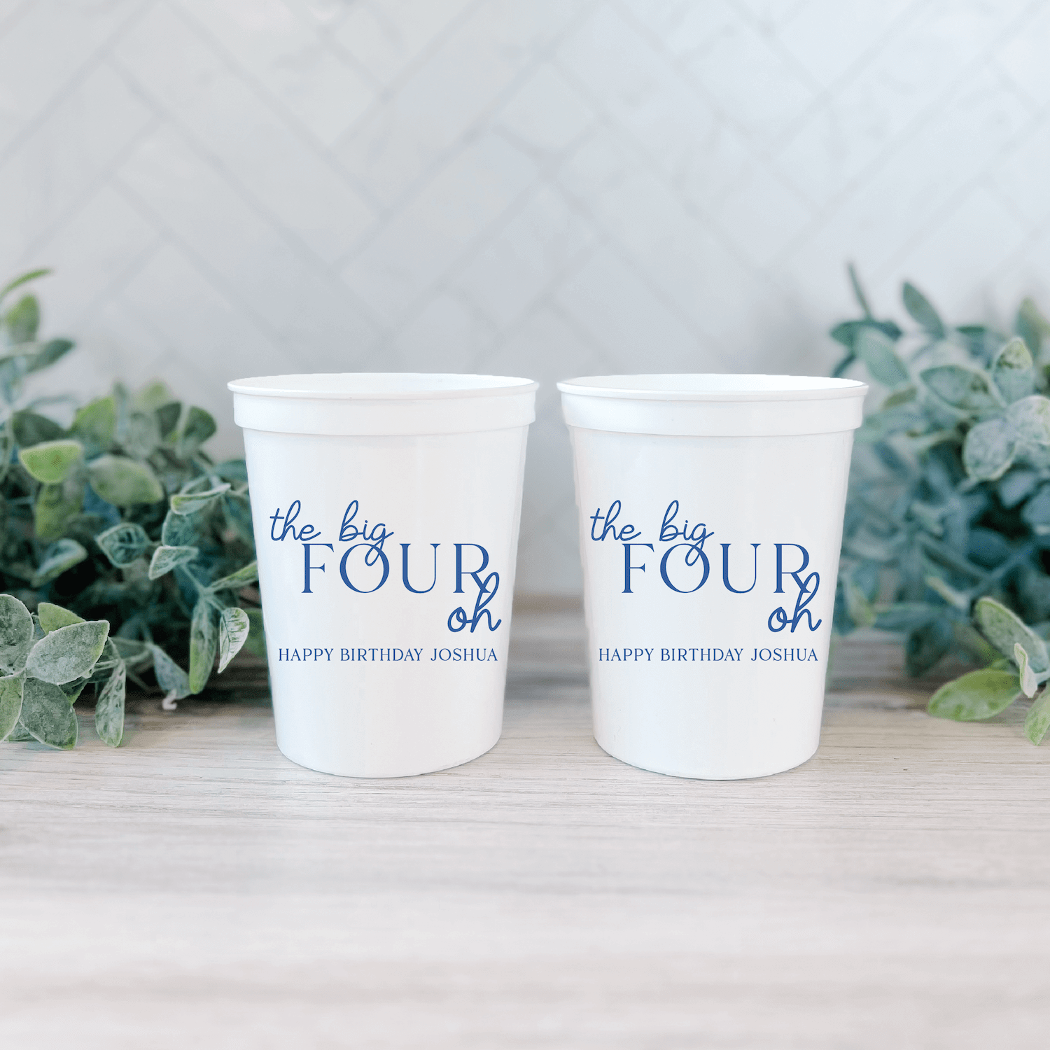 White stadium cups with "the big FOUR oh" text and "HAPPY BIRTHDAY ASHLEY" personalization, displayed with greenery