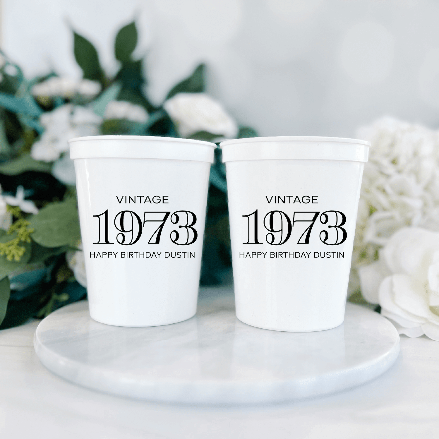 Two white stadium cups printed with "Vintage 1973" and "Happy Birthday Dustin" text, displayed on a marble coaster with greenery and white flowers.