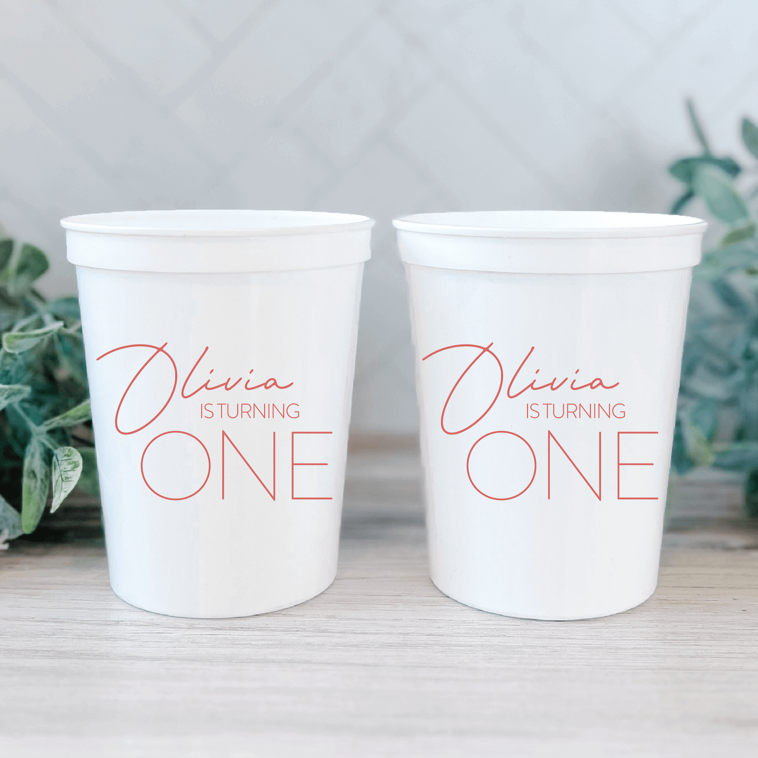 White stadium cups with pink script text reading "Olivia is Turning ONE" personalized for a first birthday party.