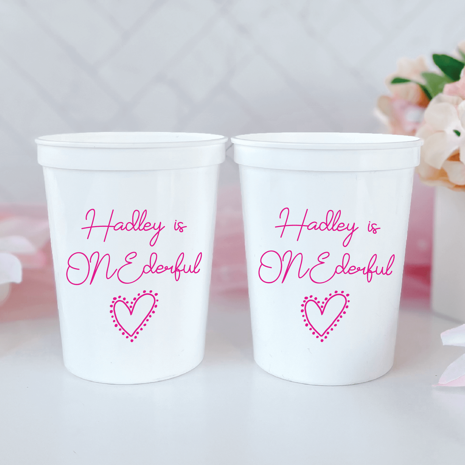 Two white stadium cups with pink text reading "Hadley is ONE derful" and a heart design, personalized for a first birthday party.