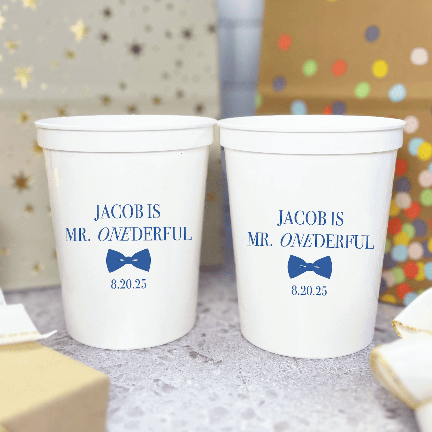 Two white stadium cups personalized with "Jacob is Mr. ONEderful" and "Jacob is Mr. ONEderful" text with blue bow tie design and date 8.20.25, displayed against colorful confetti background.