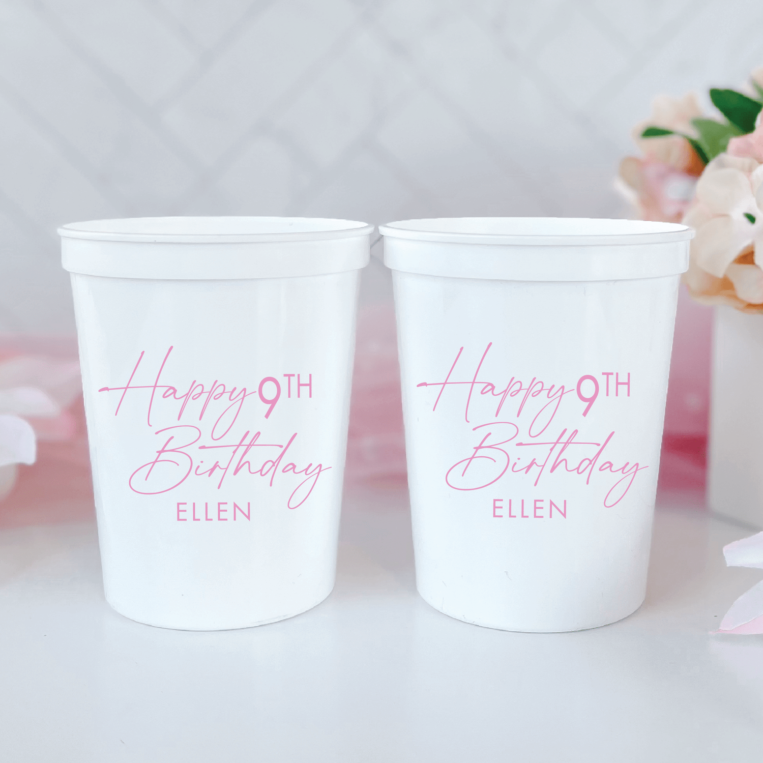 Two white stadium cups with pink script text reading "Happy 9TH Birthday ELLEN" and white flowers in background.