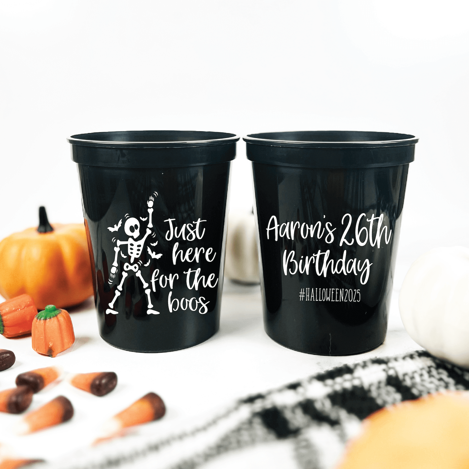 Two black stadium cups with white text, one reading "Just here for the boos" with skeleton design, the other personalized "Aaron's 26th Birthday #HalloweenBirthday," surrounded by Halloween decorations and pumpkins.