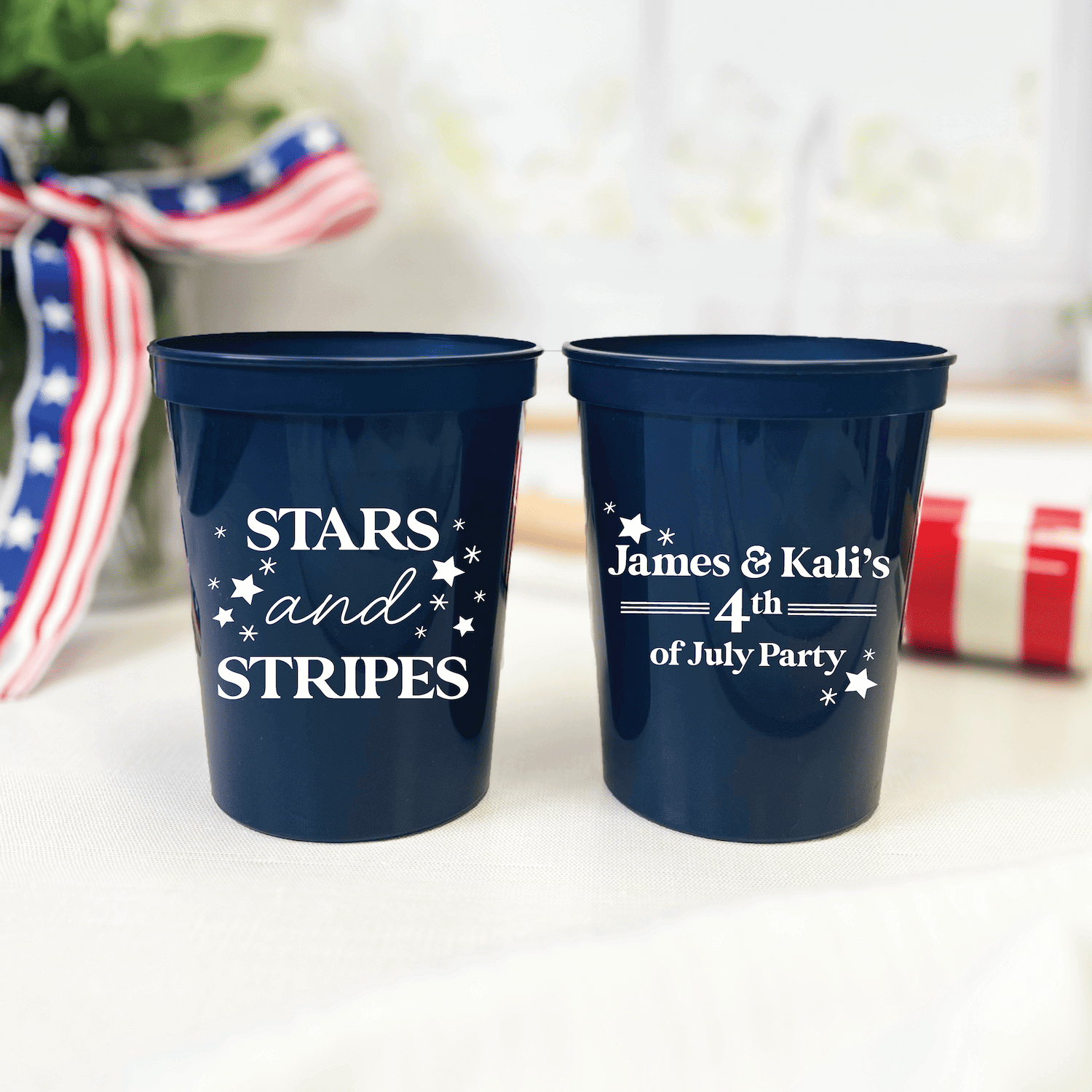 Two dark blue stadium cups with white text reading "STARS and STRIPES" and "James & Kali's 4th of July Party" with star designs, displayed with patriotic ribbon.